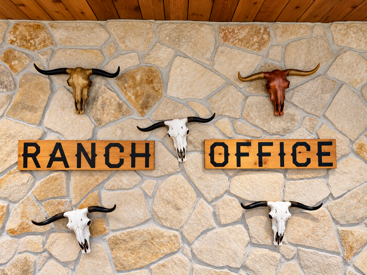 Decorative wall with four animal skulls with horns mounted above two wooden signs, one reading 'Ranch' and the other 'Office', on a stone wall with a wooden ceiling.