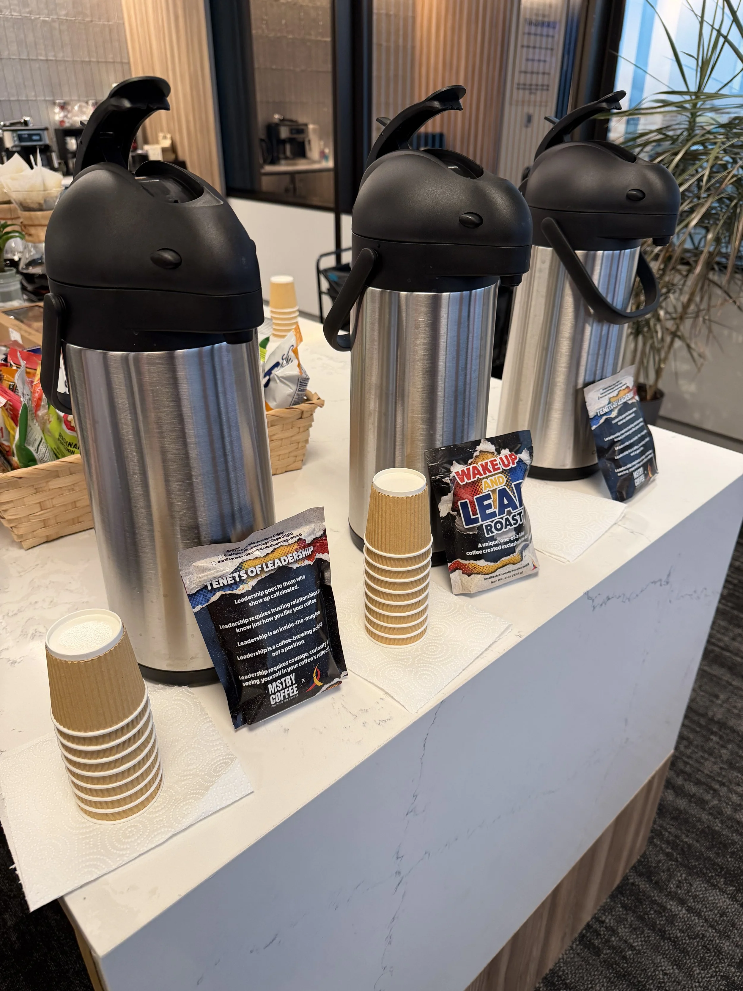 Three large stainless steel coffee dispensers with black lids and handles on a white countertop. There are paper cups stacked in front of the dispensers, and packets of coffee labeled "Wake Up and Leaf Roast." A small sign with info about leadership and coffee is also on the counter. In the background, there are baskets with snack items and a coffee station.