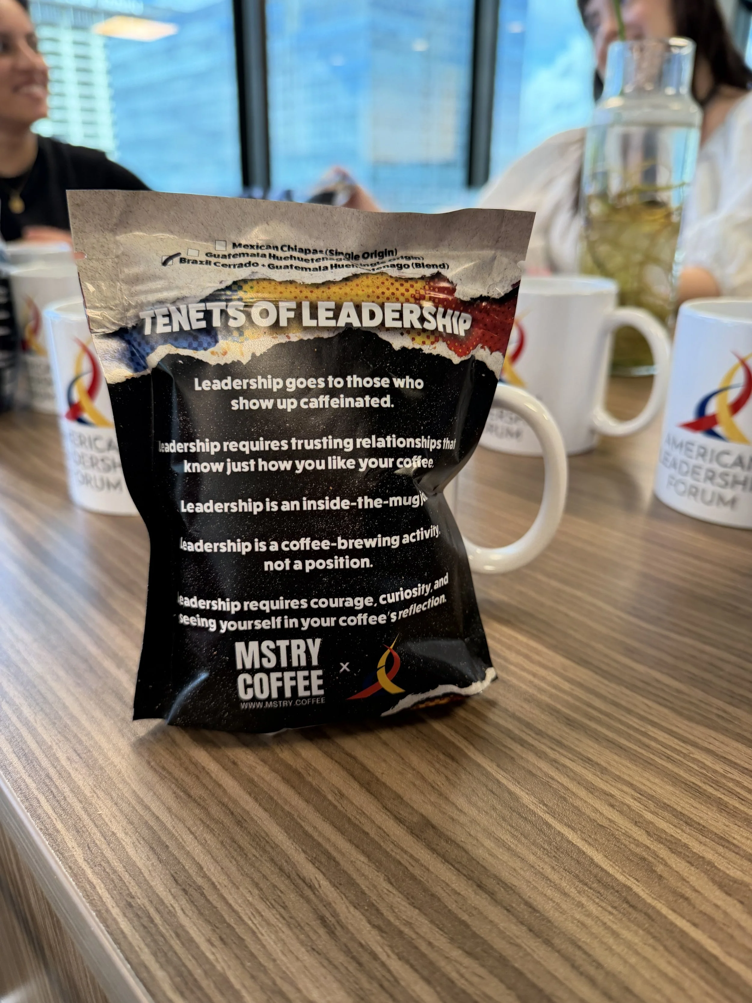 A torn packet of coffee placed on a wooden table in front of several mugs and a glass of iced beverage, with people sitting around the table in a bright room with large windows.