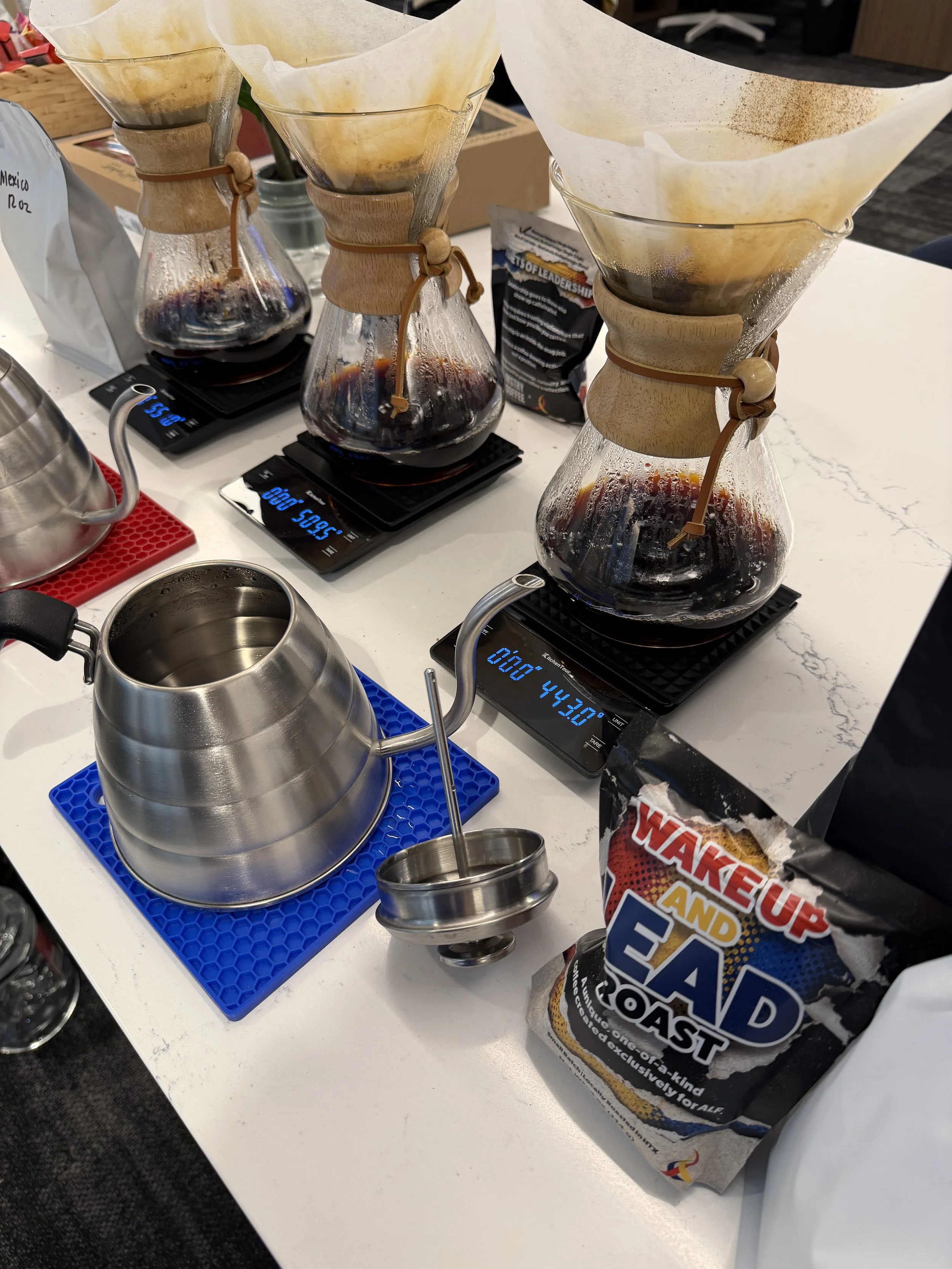 Three Chemex coffee makers with paper filters and coffee inside, placed on digital scale counters, on a white table in a workspace. An empty metal gooseneck kettle and coffee bags are also visible.