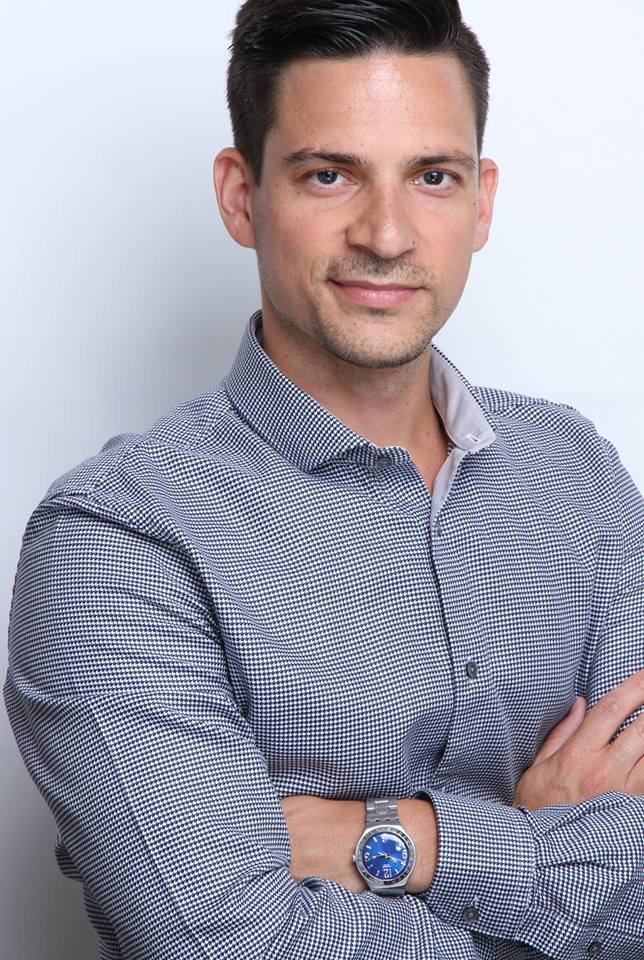Man in light checkered shirt with neatly styled hair and trimmed circle beard, slightly smiling, standing with arms crossed and wearing silver watch with blue on it.