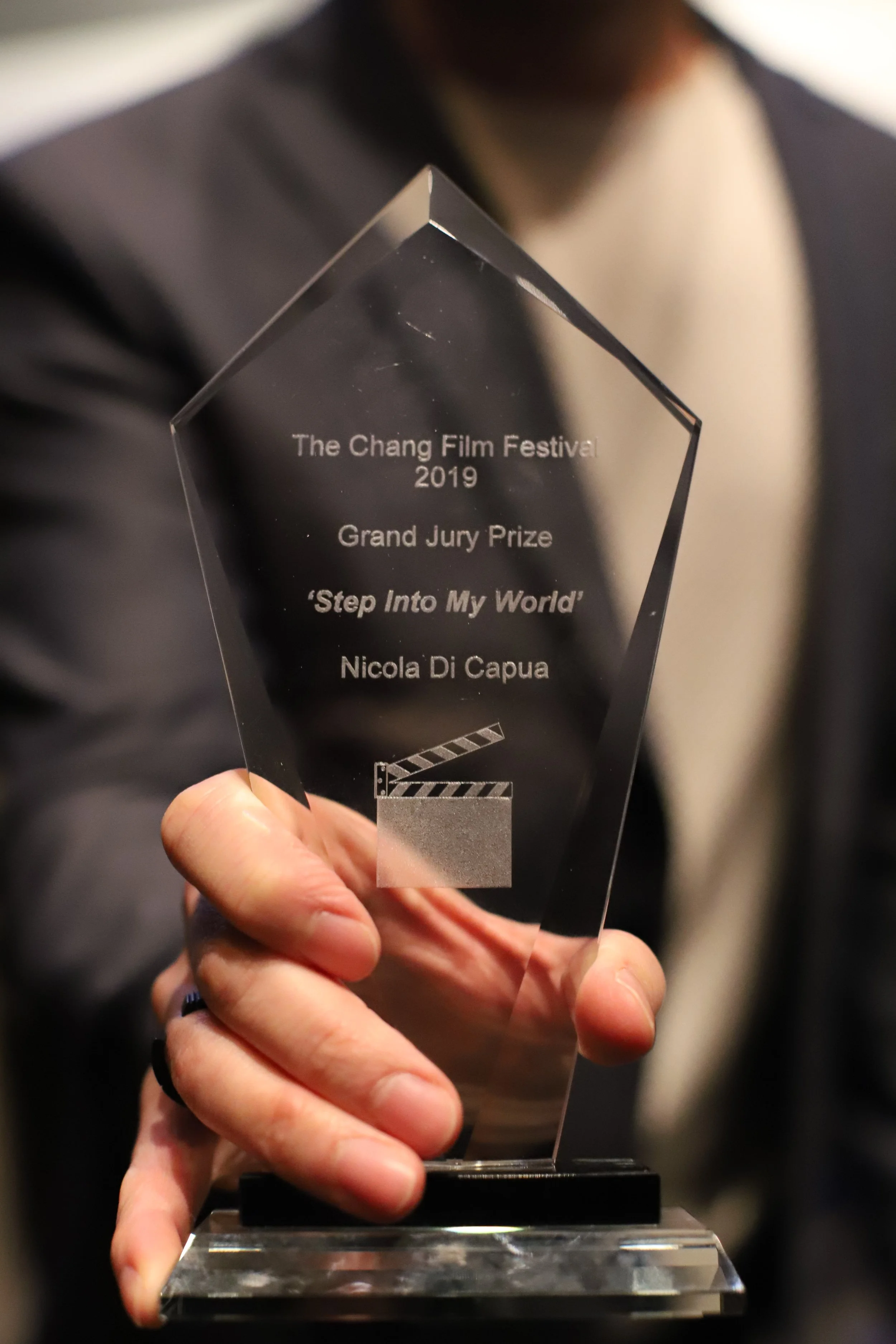 Close-up of transparent glass film award in tall diamond shape held in hand. Background shows blurred upper body. Engraved with text: The Chang Film Festival 2019, Grand Jury Prize, 'Step Into My World,' Nicola Di Capua, with film clapperboard icon.