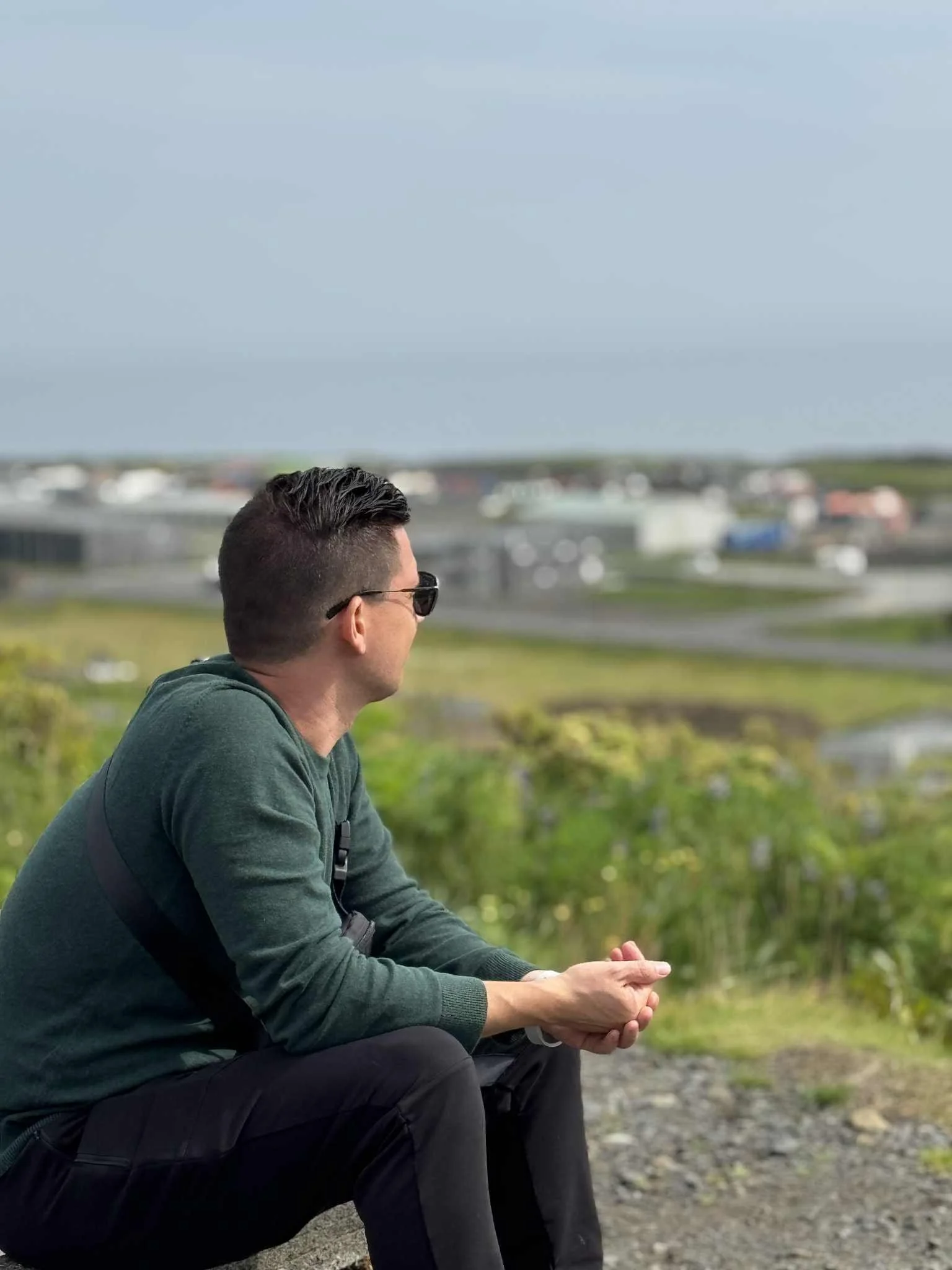 Man wearing dark sunglasses, green long-sleeve shirt, and blank pants sits on rock outdoors on grassy hill with hand clasped, looking into distance toward town in the background under blue sky.