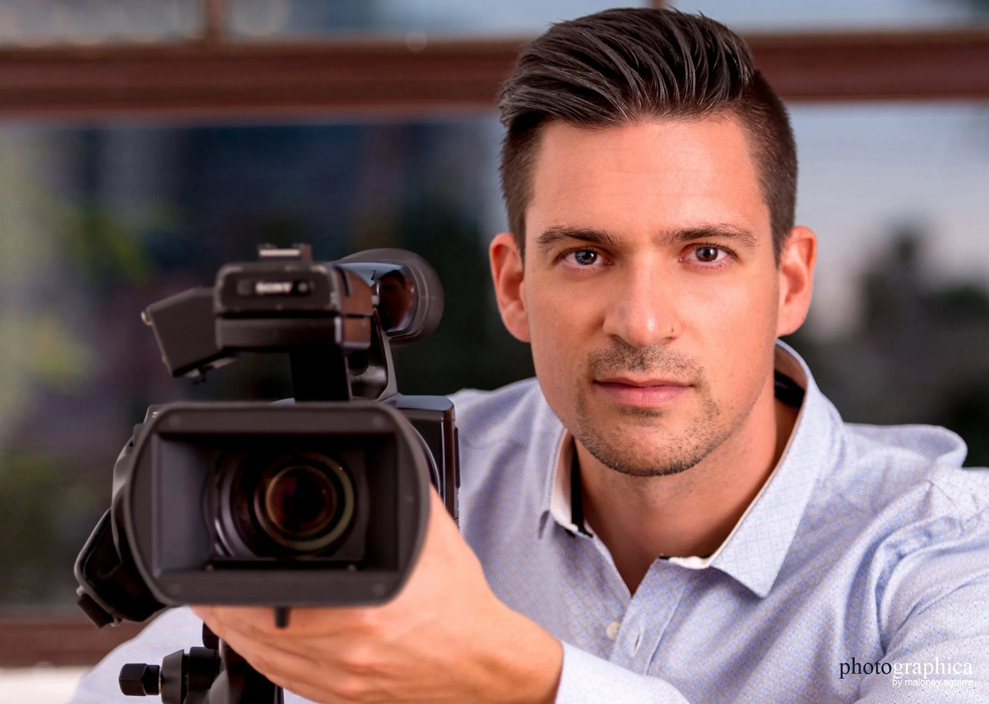 Close-up of man in light blue collared shirt with neatly trimmed circle beard, slighly smiling, looking directly at the camera while holding professional video camera with his left hand.