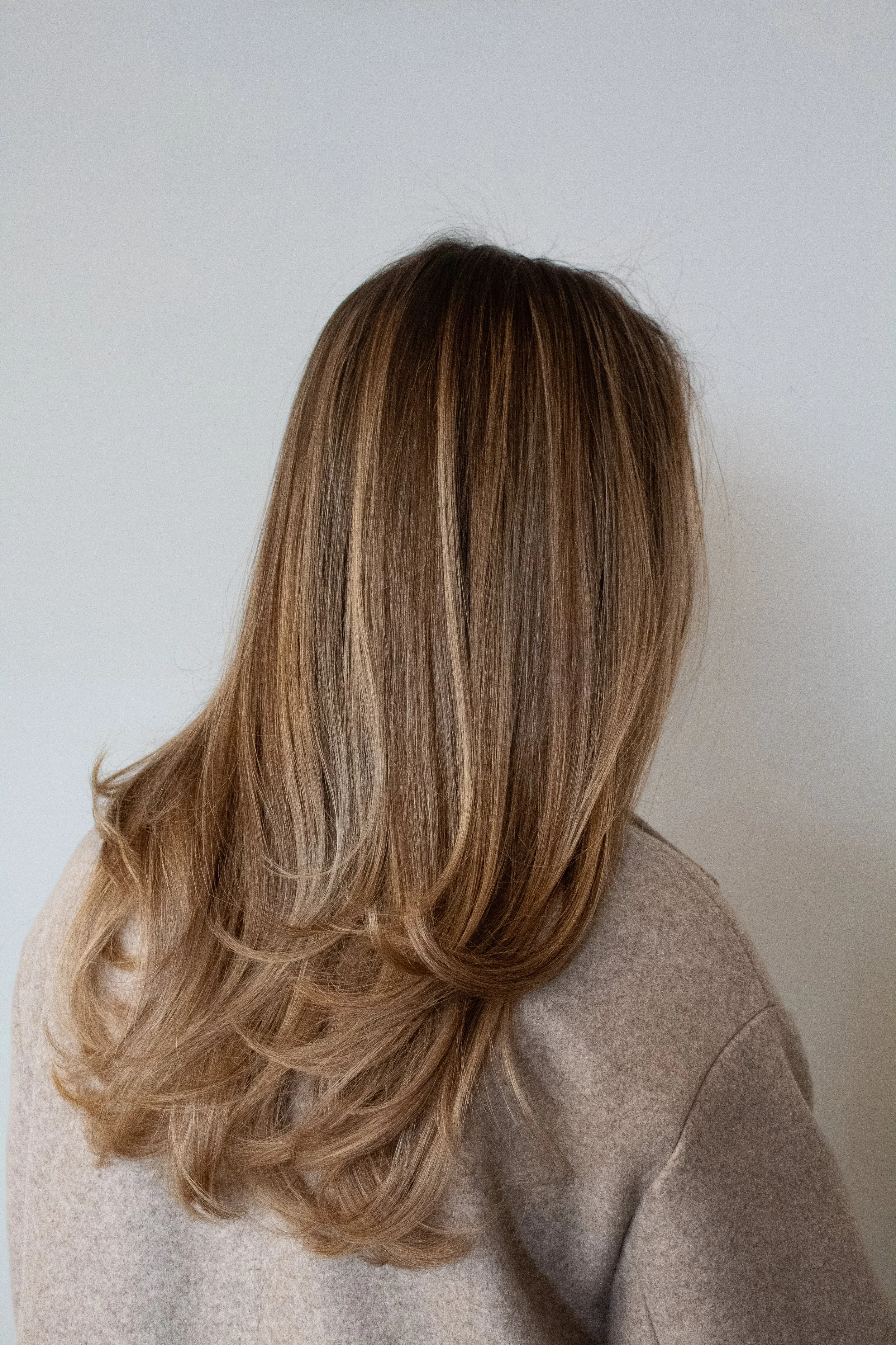 Side view of a client with a blonde blowout by stylist Traci against a white backdrop. The client is wearing a beige coat.