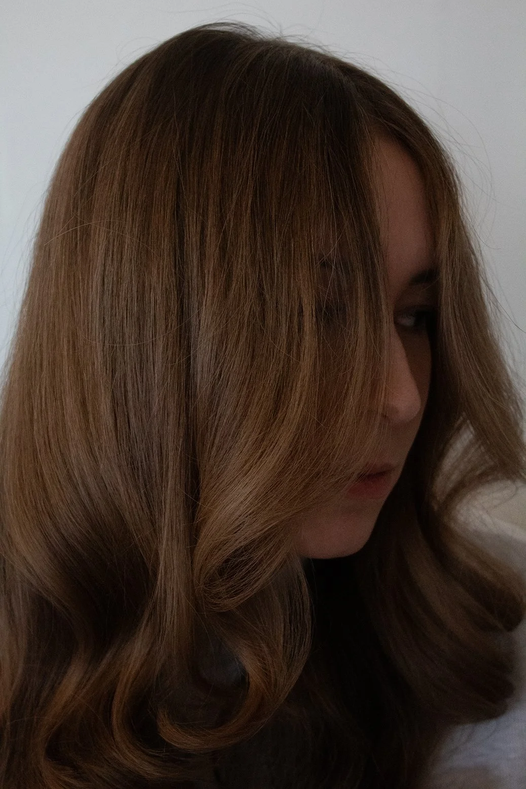 Side view of client with wavy, brunette hair that partially covers her face as she looks downward against a white backdrop.