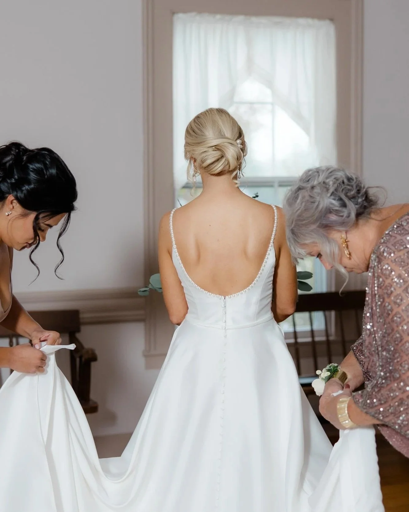 Back view of a bride in her white gown, her hair twisted into an updo with two women holding up the ends of her train
