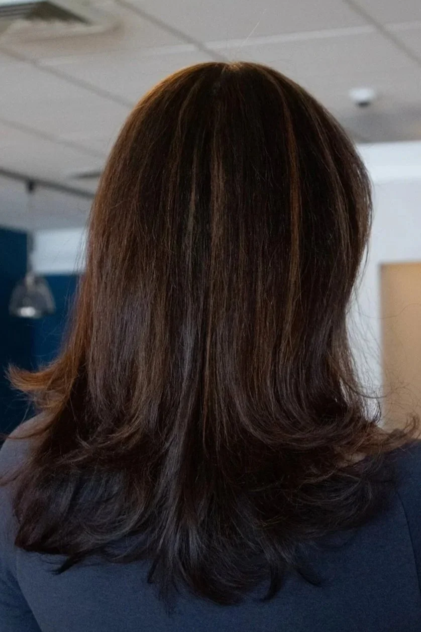 Back view of a client with shoulder-length brown hair with subtle highlights against a salon backdrop. Hair is blown out and styled.