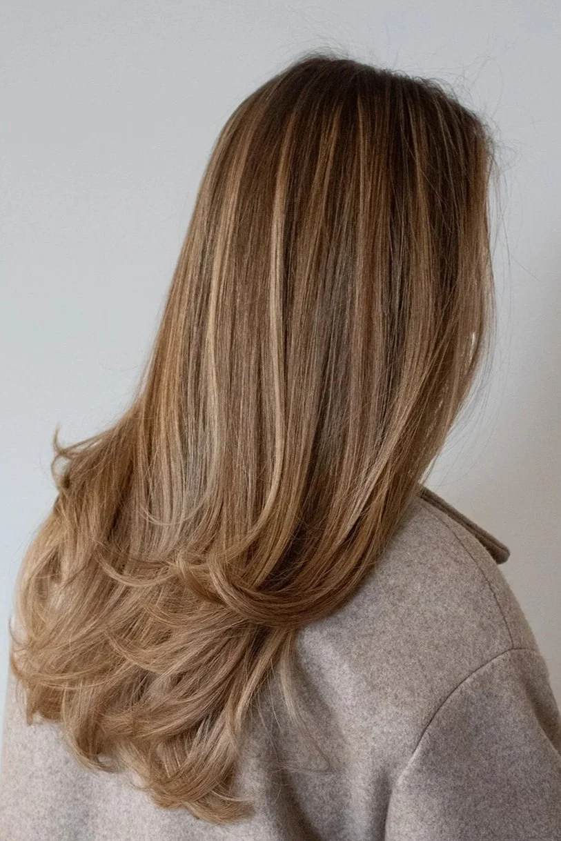 Side view of a client with a blonde blowout by stylist Traci against a white backdrop. The client is wearing a beige coat.