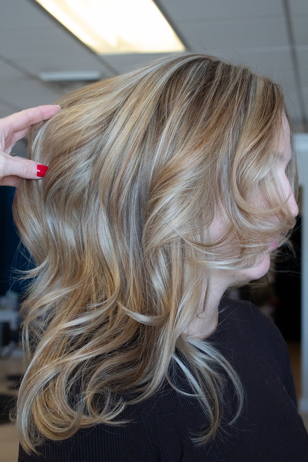 Side view shot of client with fresh blonde highlights, with stylist's hand fanning out hair on the left side, against a salon backdrop.
