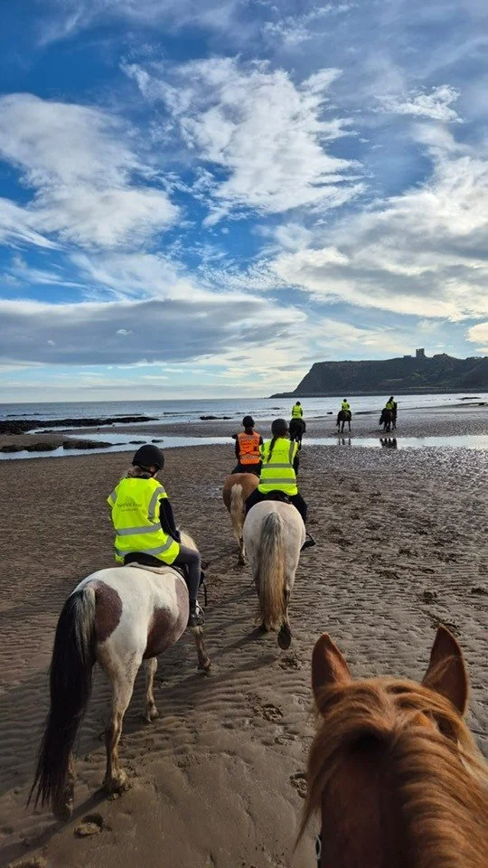 People riding horses along a sandy beach under a partly cloudy sky.
