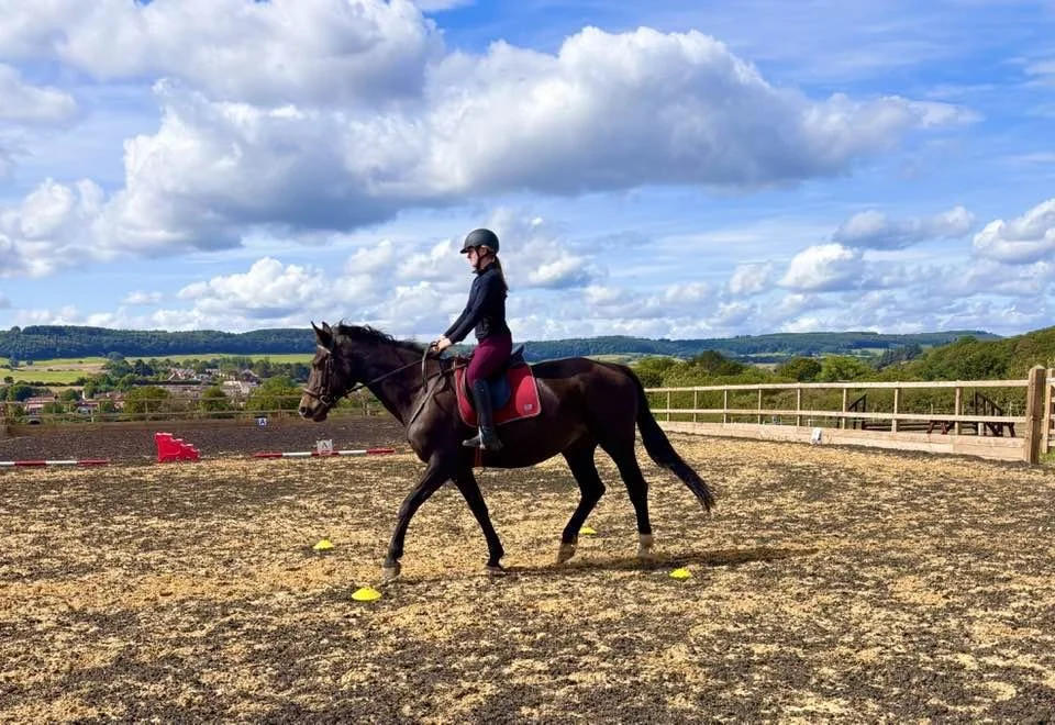 A woman riding a black horse in an outdoor riding arena on a sunny day with scattered clouds.