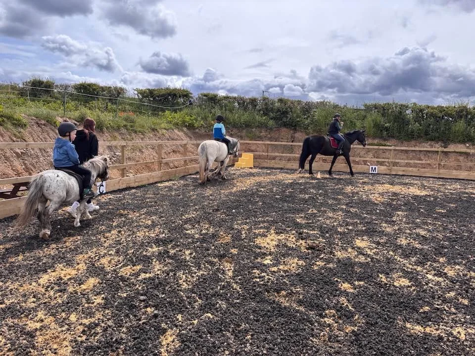 Three children riding ponies in an outdoor riding arena on a cloudy day, with two adults supervising.