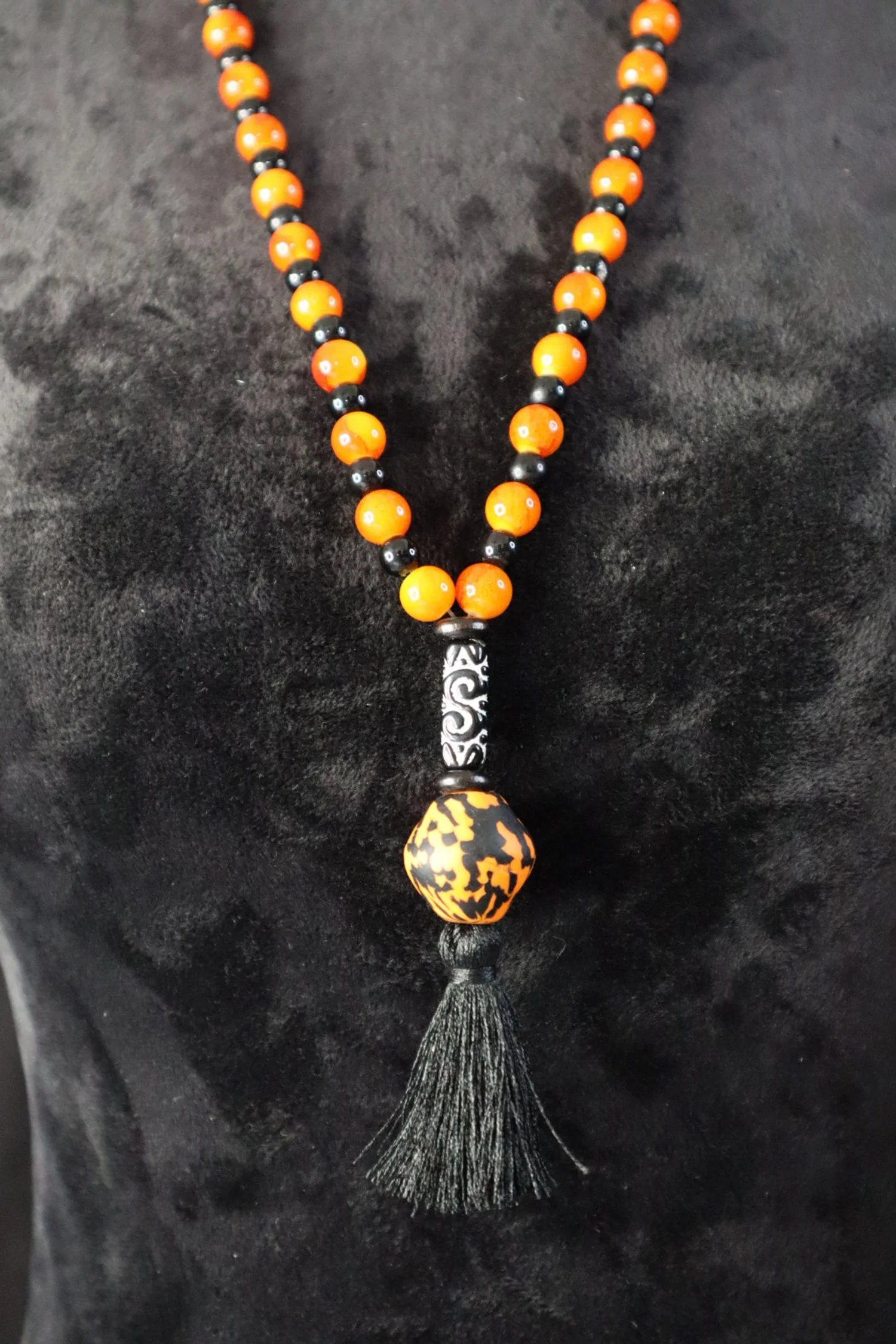 A beaded necklace with orange and black beads, featuring a decorative black and white segment, an orange and black patterned bead, and a black tassel.