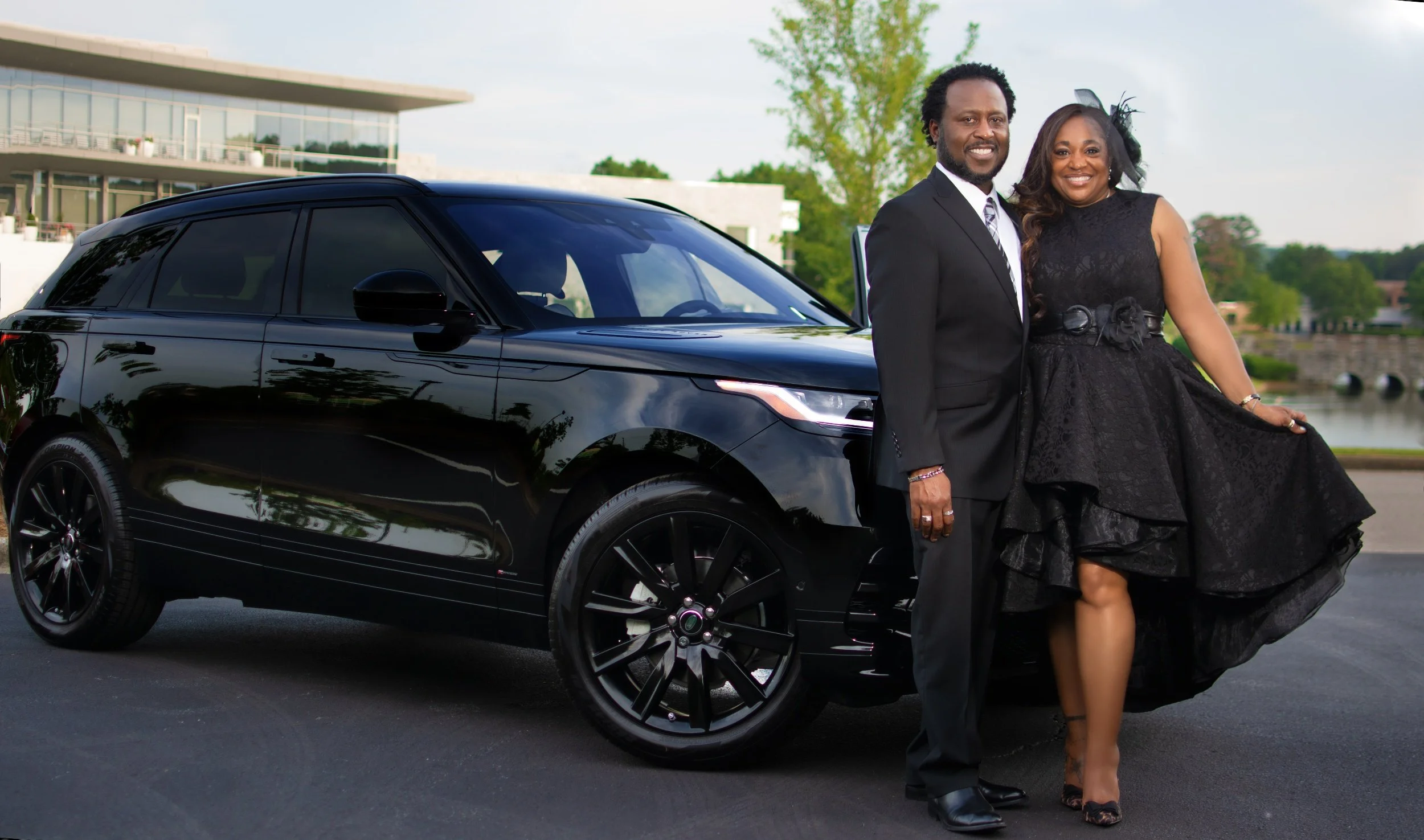 A happy couple dressed in formal black attire standing next to a black luxury SUV outdoors near a waterfront.
