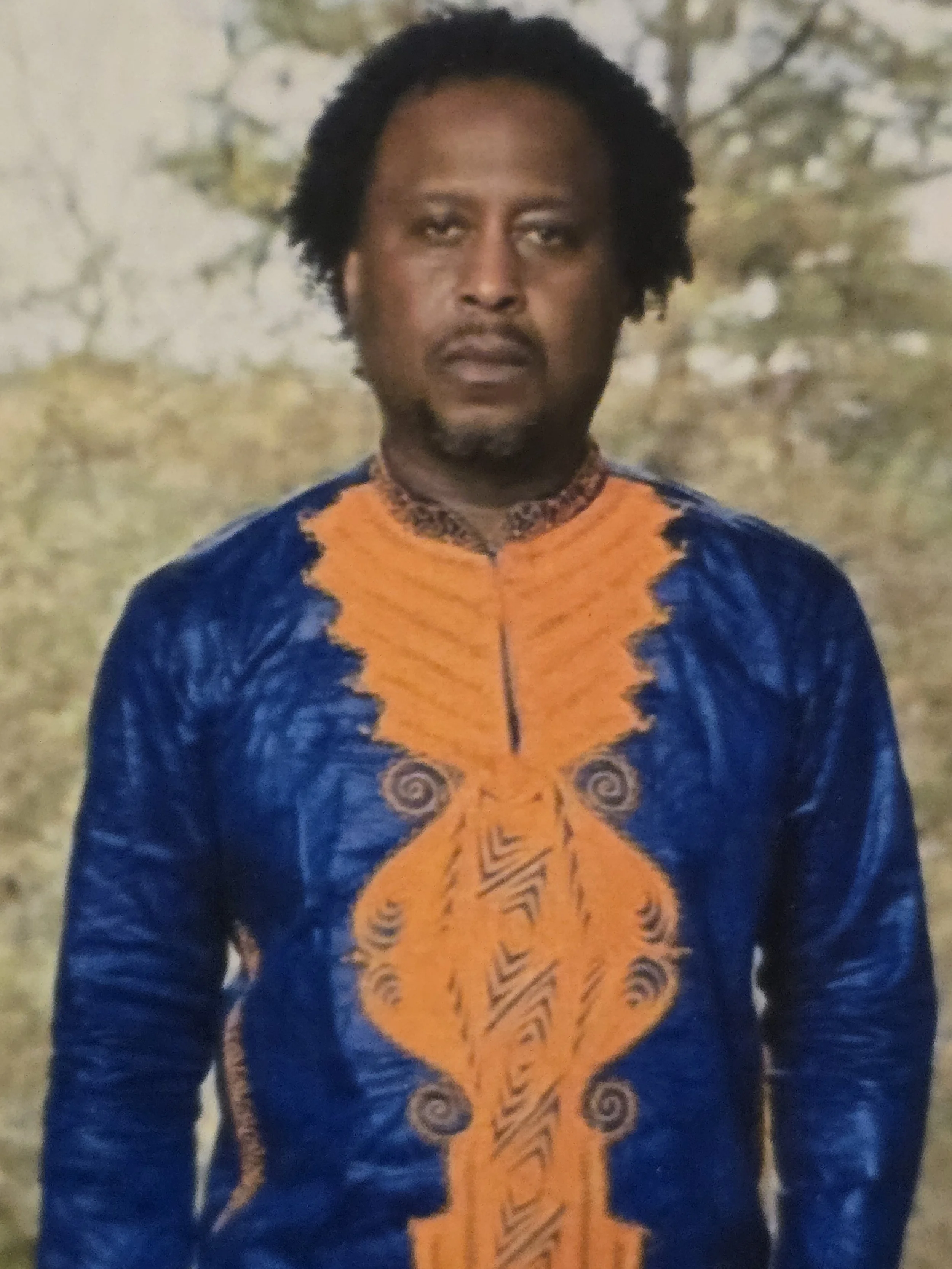 A man with dark skin and curly hair wearing a colorful traditional African shirt with orange and blue patterns, standing outdoors with a blurred background of trees.
