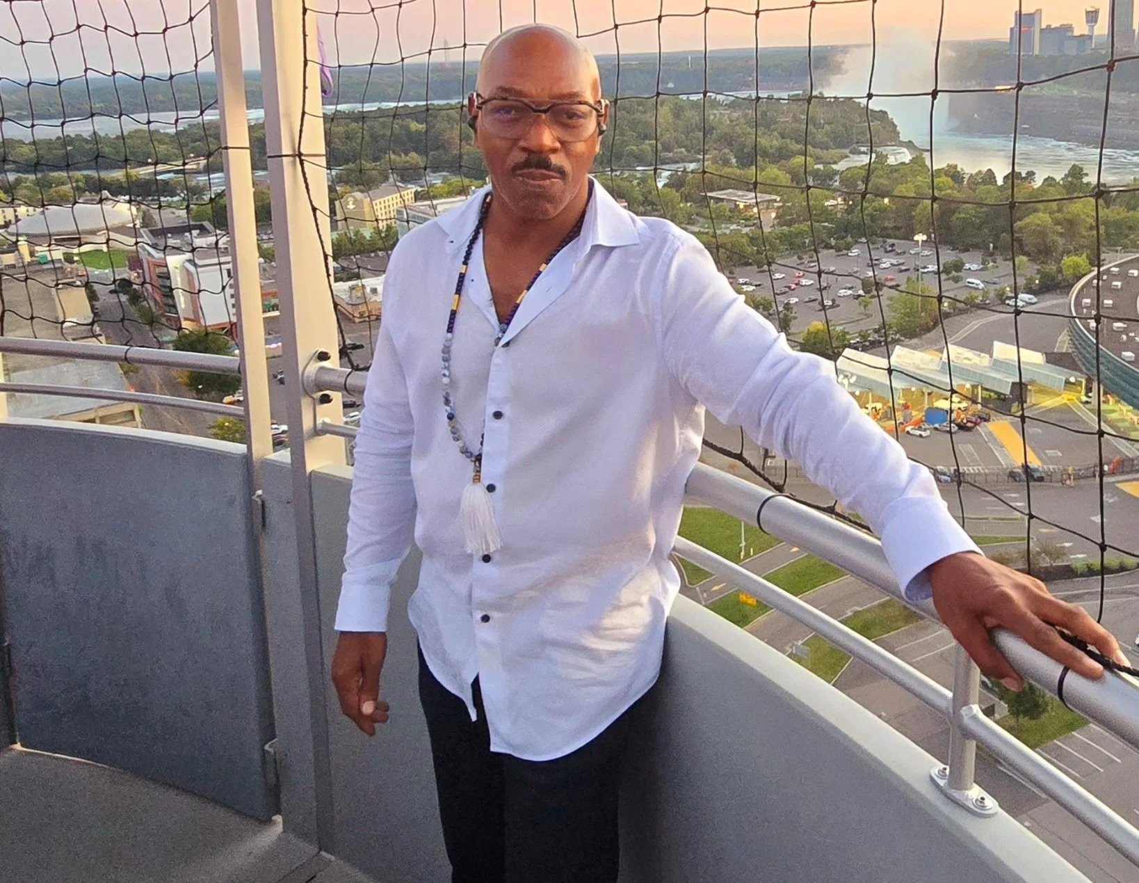 A man with glasses and a white shirt stands on a rooftop observation deck with safety netting, overlooking a cityscape at sunset, including a river, parks, and parking lots.
