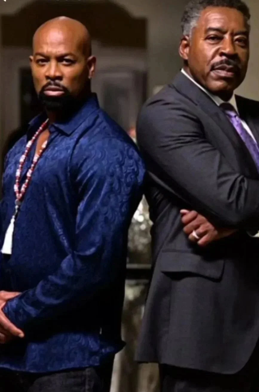 Two men standing back to back in a serious pose, one in a blue patterned shirt with bead necklaces, and the other in a dark suit with arms crossed.
