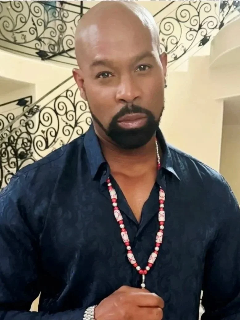 Darrin Henson, wearing a dark blue shirt, a beaded necklace, and a watch, standing indoors in front of a decorative wrought iron railing.