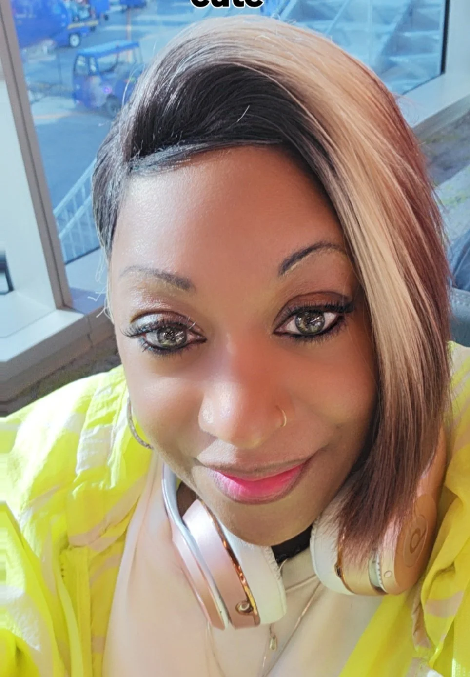 A woman with shoulder-length, two-toned hair, featuring black and blonde sections, smiling. She has a nose ring, wears makeup, and has headphones around her neck. She is wearing a yellow jacket and is inside a building near a window, with cars visibl