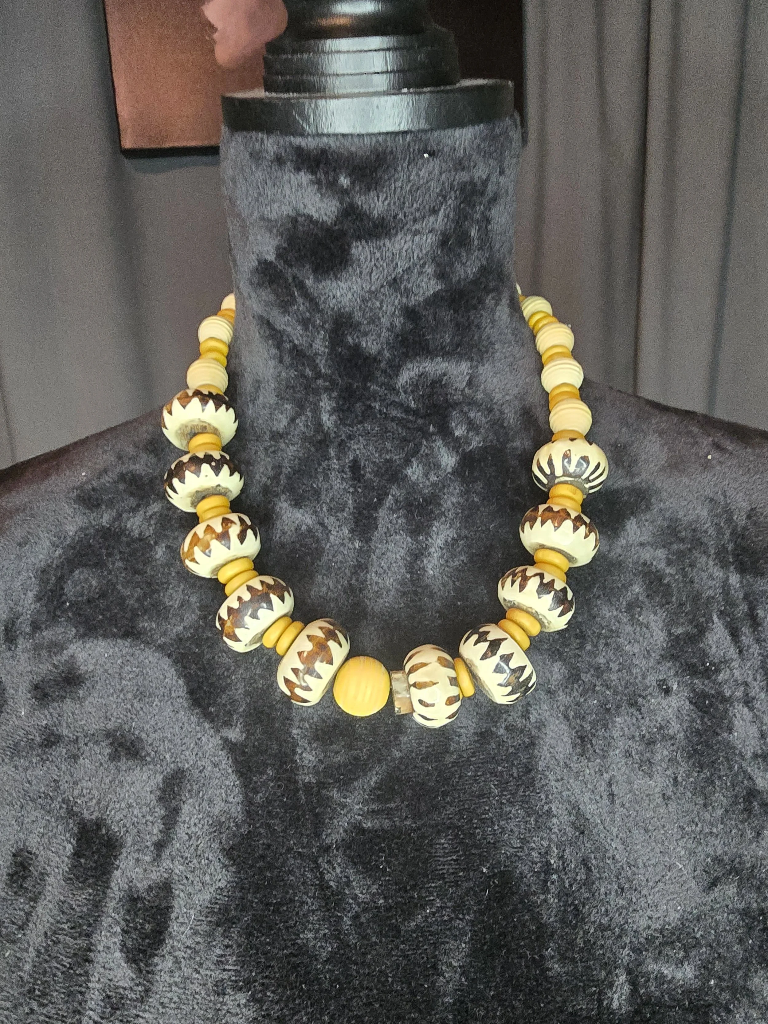 Beaded necklace displayed on a black velvet bust, featuring large ivory and brown patterned beads separated by small yellow spacer beads.