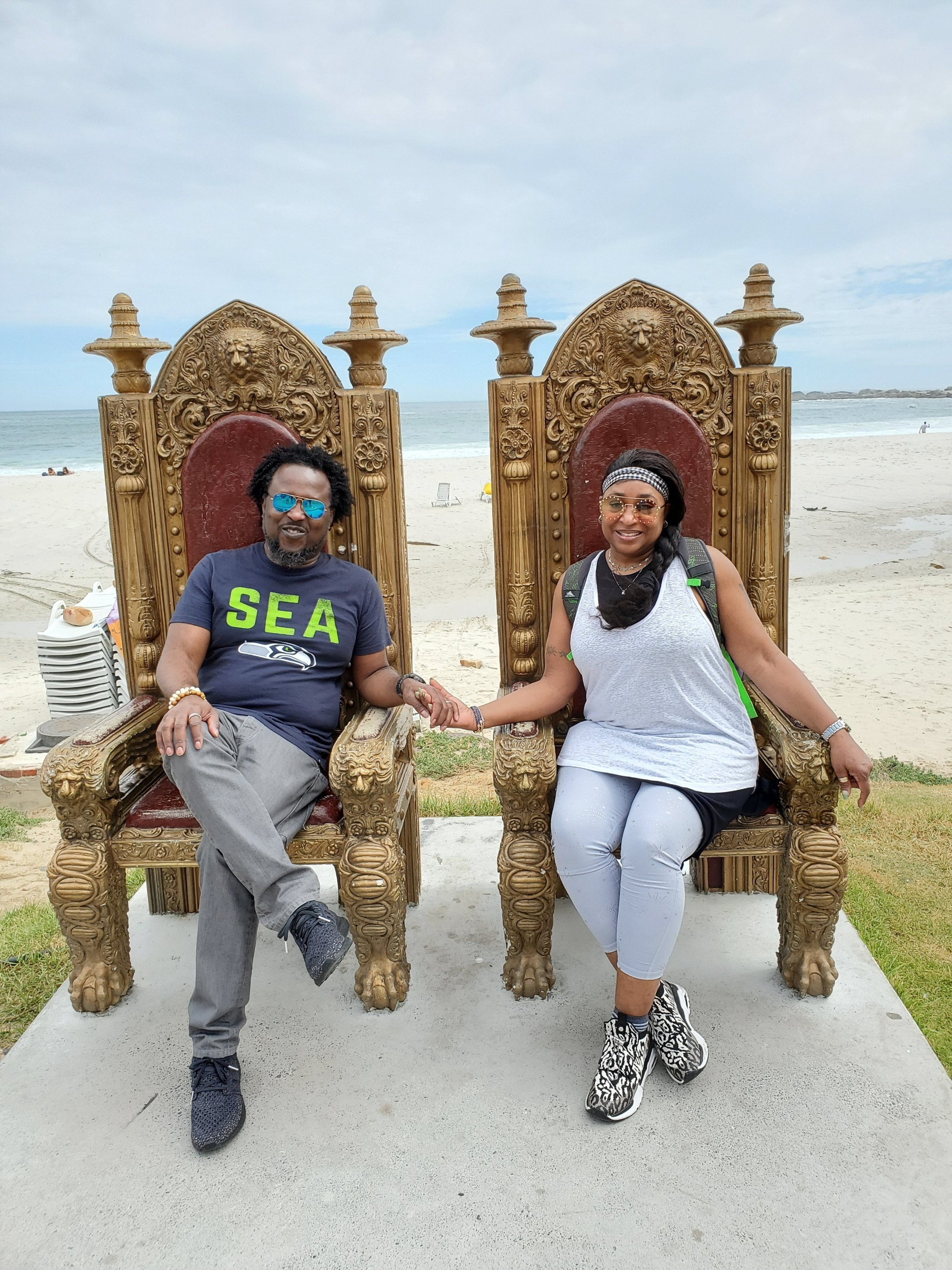 A man and woman sitting on large ornate wooden chairs on the beach, holding hands and smiling. The ocean and cloudy sky are in the background.