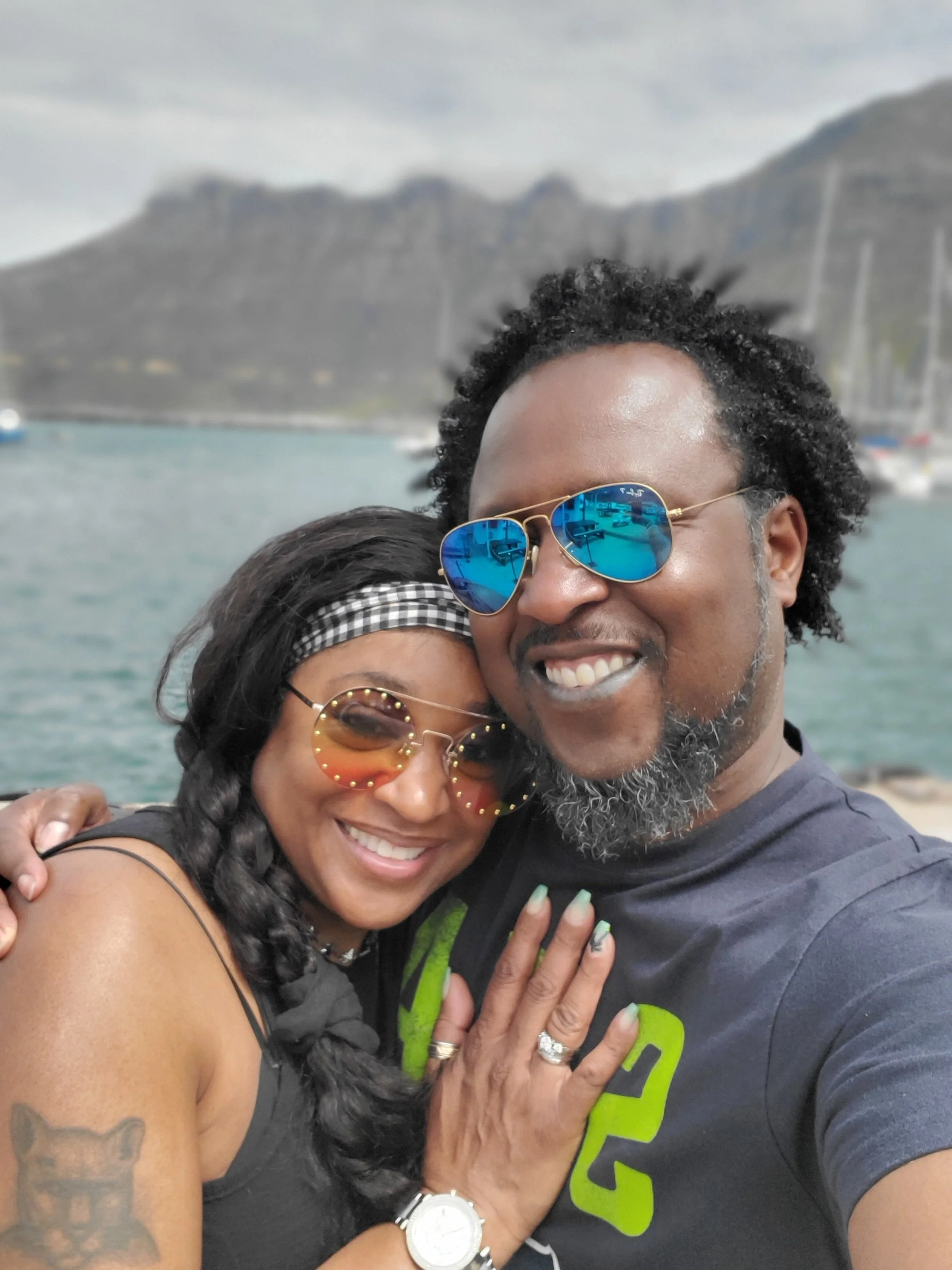 A smiling couple wearing sunglasses, embracing outdoors near the water with mountains and boats in the background.
