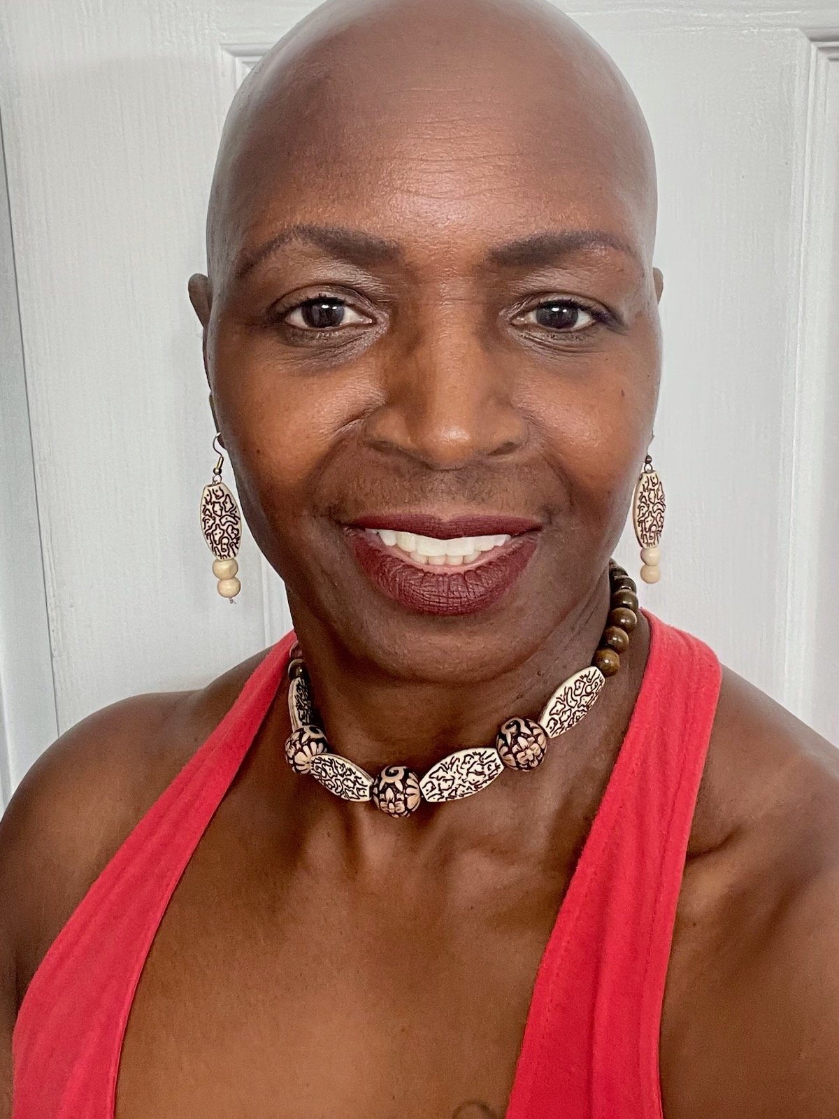 A woman with a shaved head smiling, wearing a red sleeveless top, with intricate black and white jewelry including a necklace and earrings, standing in front of a white door.