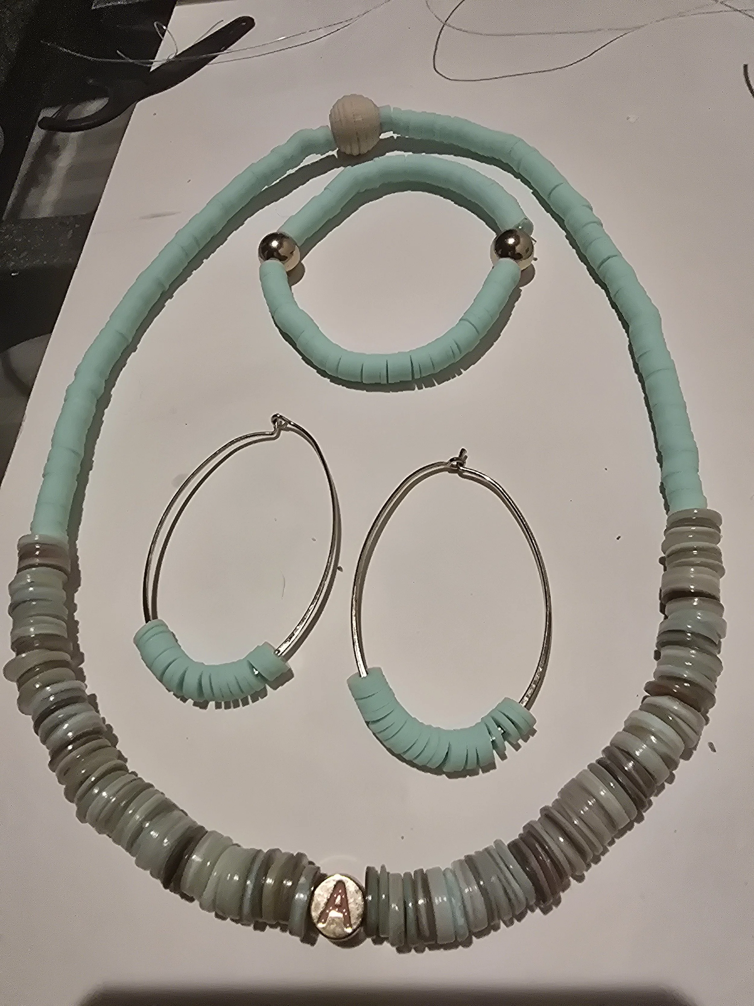 A matching jewelry set consisting of a beaded necklace with an initial 'A', a matching bracelet, and a pair of hoop earrings, all featuring light blue, gray, and beige beads.