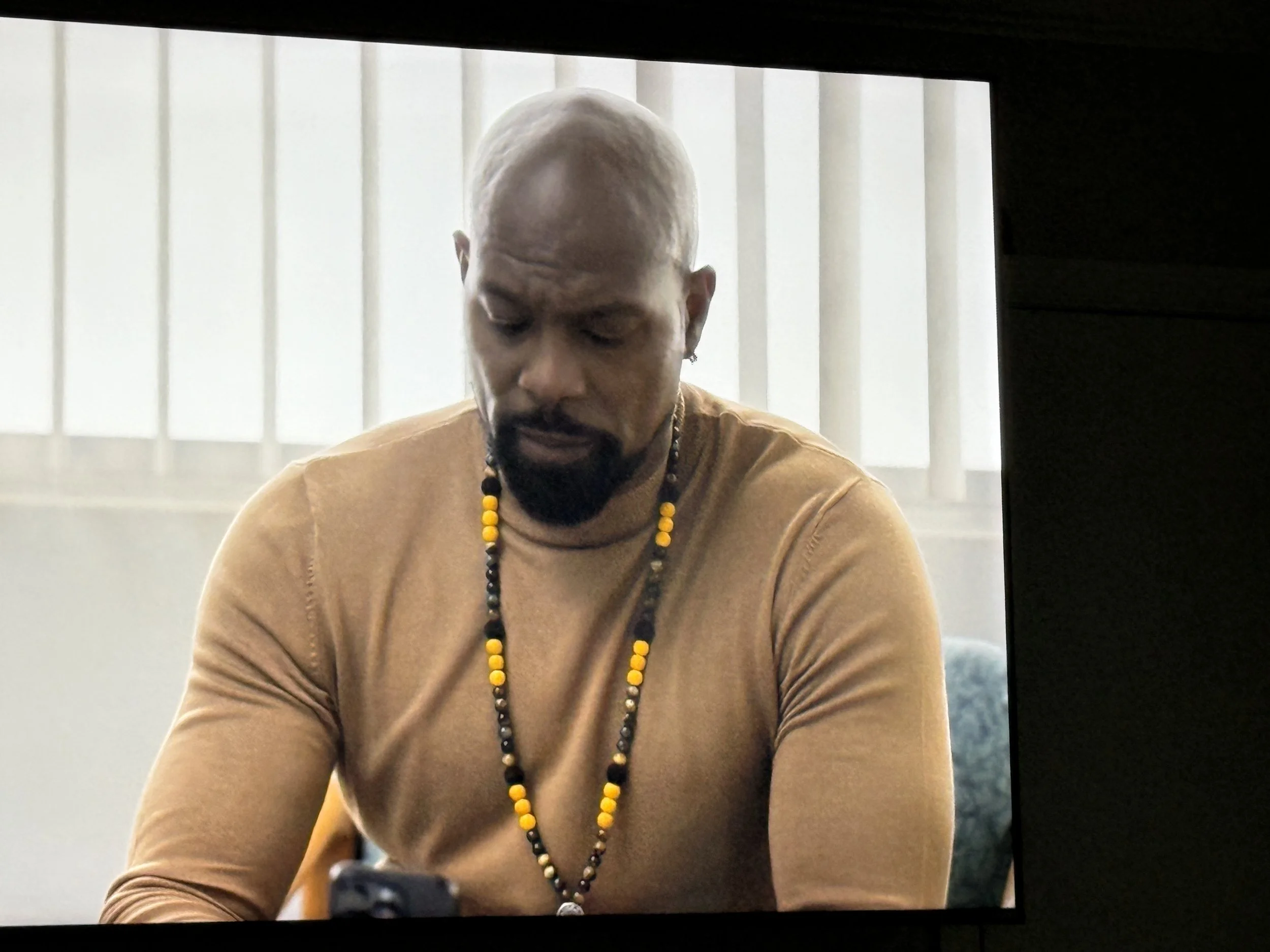 Darrin Henson, wearing a tan long-sleeve shirt and beaded necklace, looking down while sitting in front of a white wall with vertical blinds.