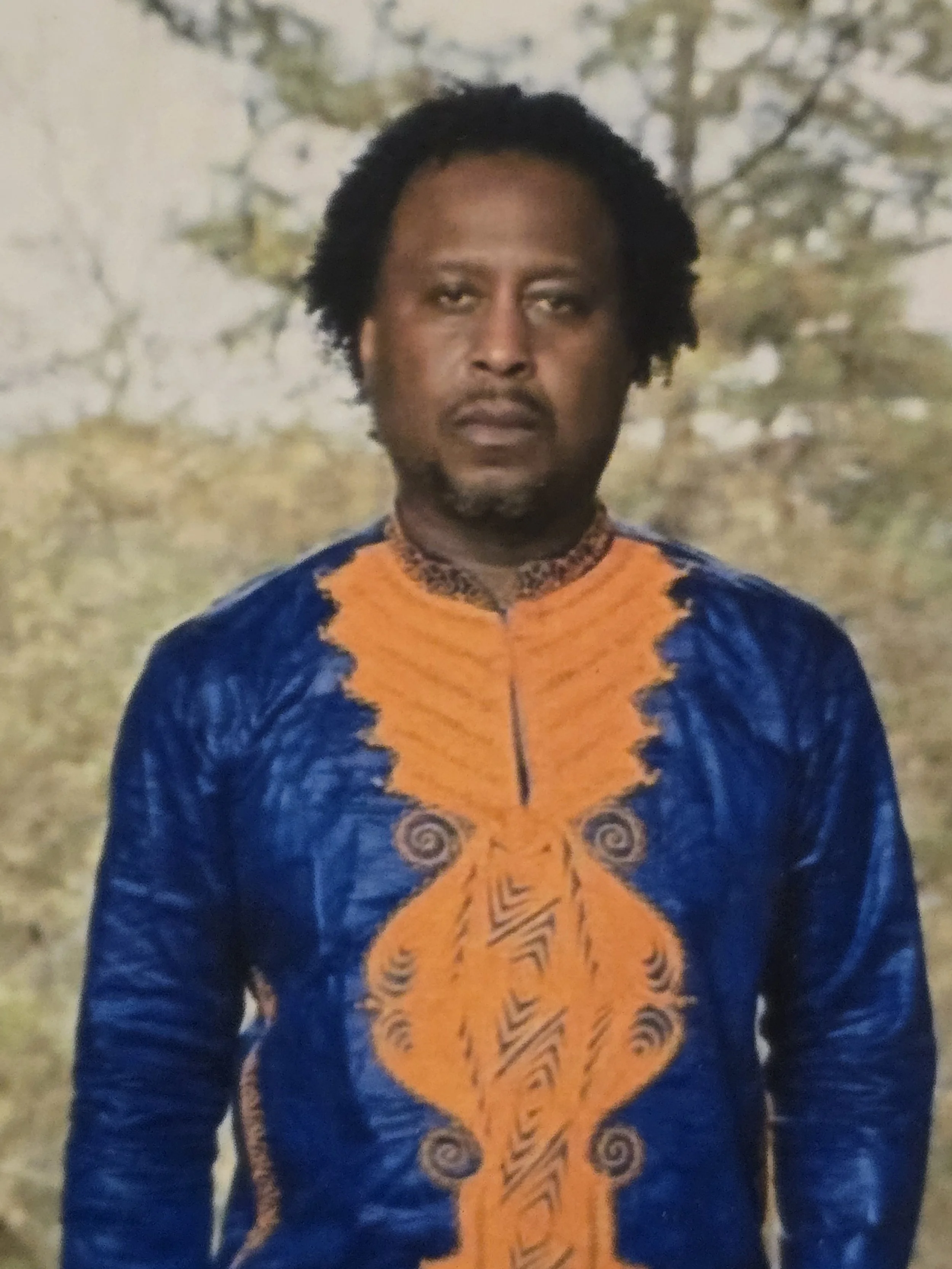 Portrait of an African American man with curly hair wearing a colorful, patterned, African-style shirt outdoors with trees in the background.