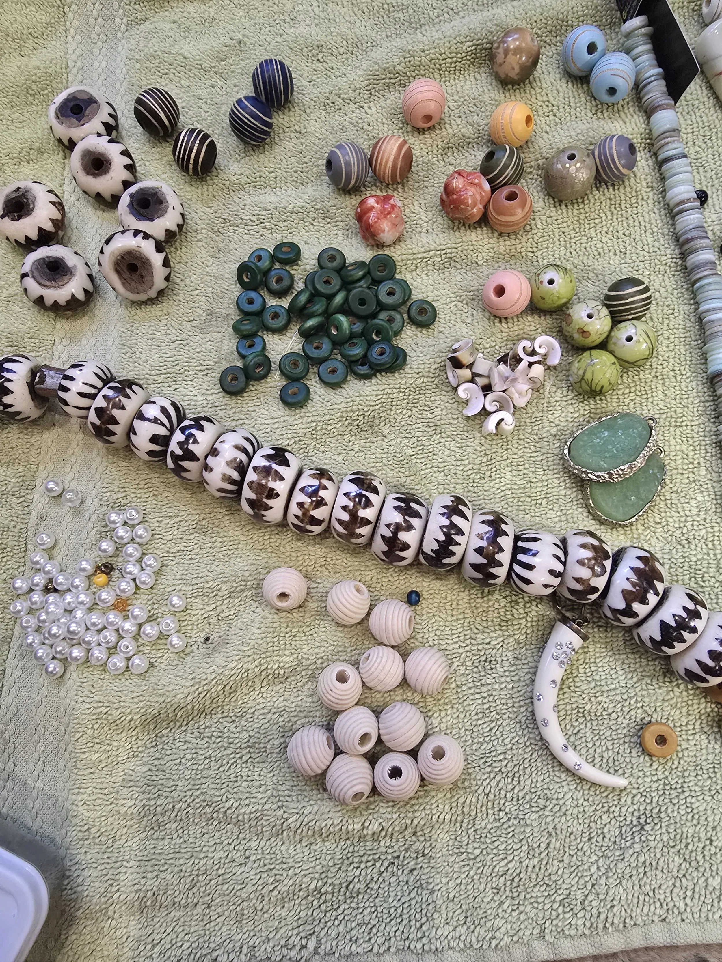 Assorted jewelry beads and findings laid out on a green towel, including black and white patterned beads, pearl-like beads, green and pink beads, and other decorative elements for jewelry making.