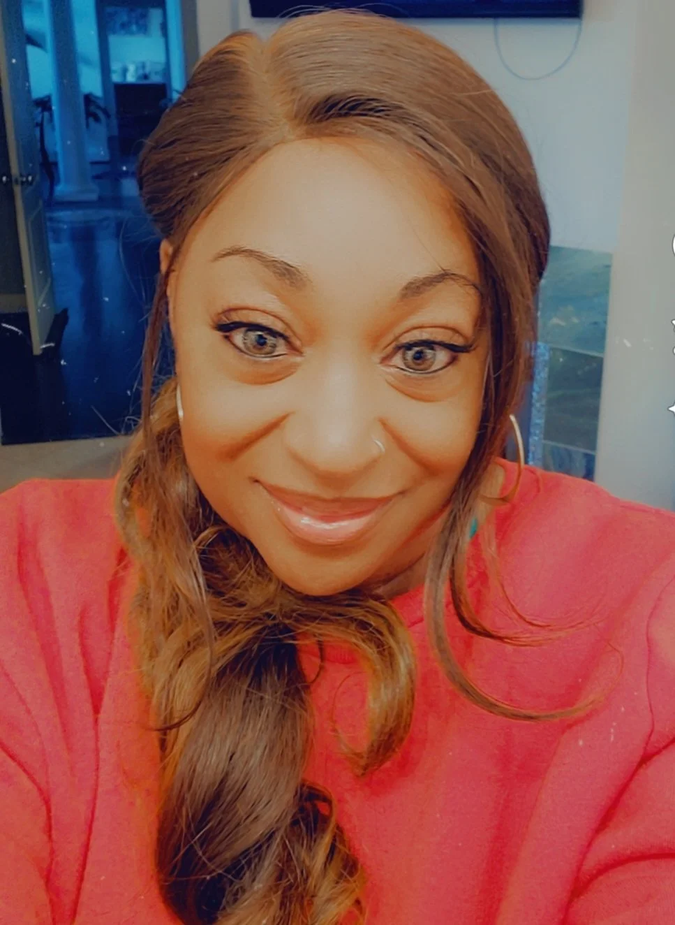 A woman taking a selfie, smiling, with long wavy brown hair, wearing a red top, in an indoor setting with a blurred background.