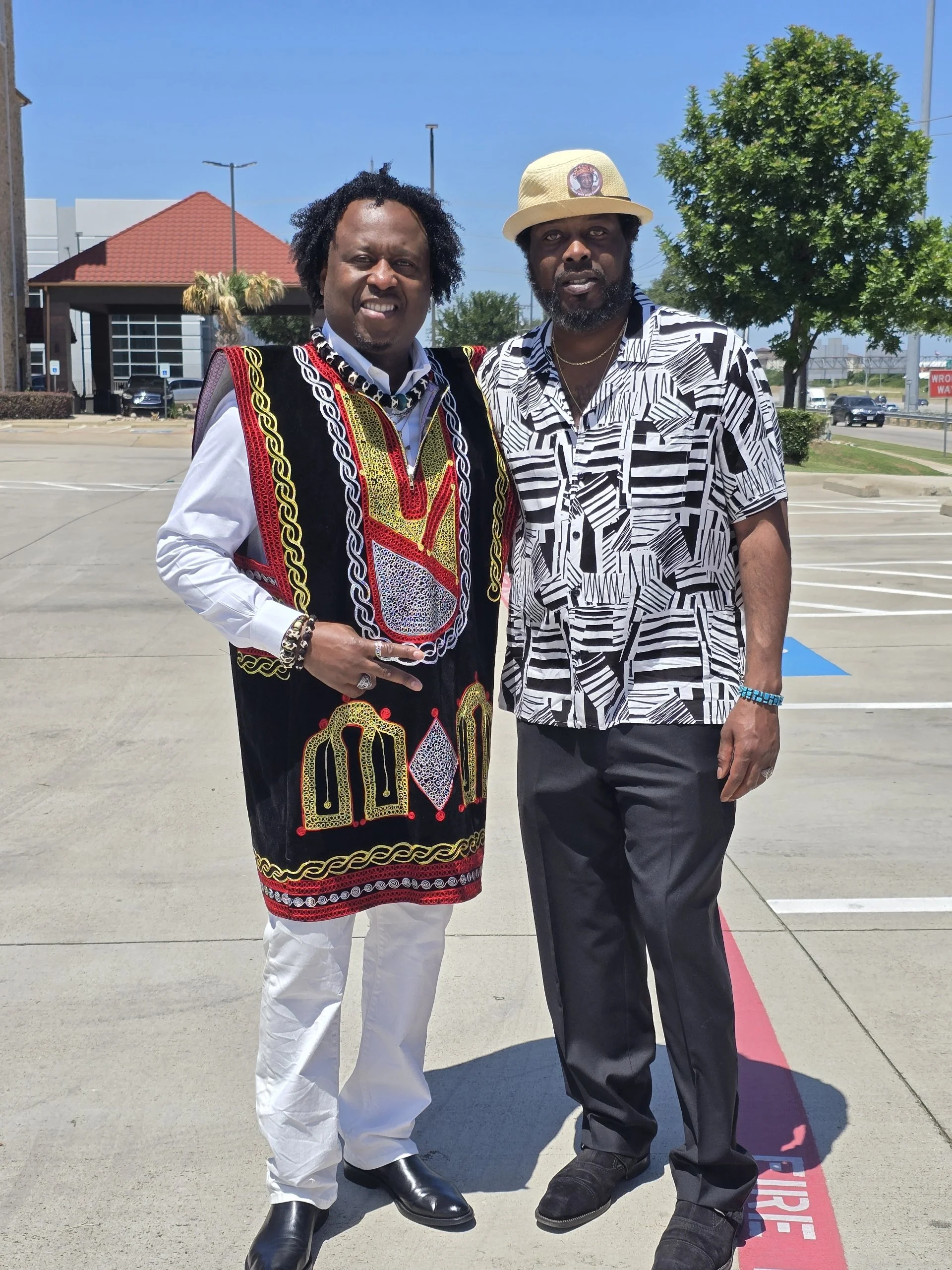 Two men standing outdoors in a parking lot on a sunny day, one dressed in vibrant African attire and the other in a black and white patterned shirt with a hat.