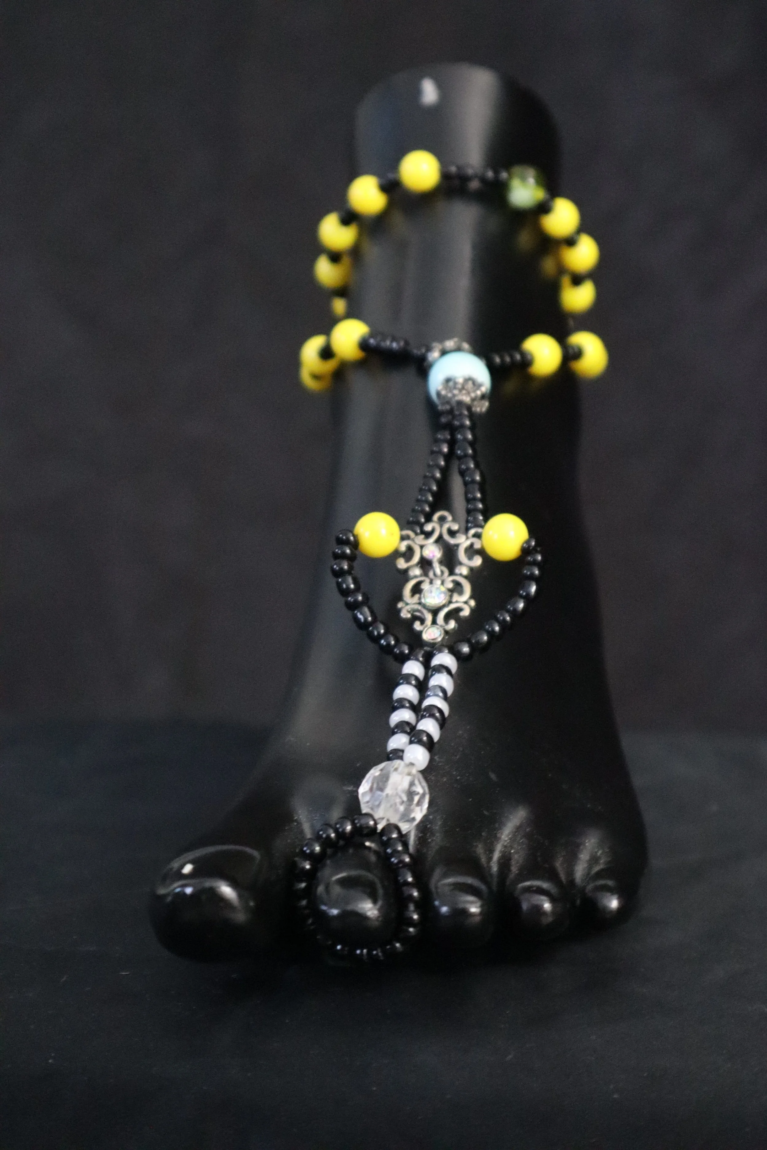 A black display foot with a beaded necklace draped over it. The necklace has a mix of black, yellow, white, and clear beads, with some metallic and decorative accents.