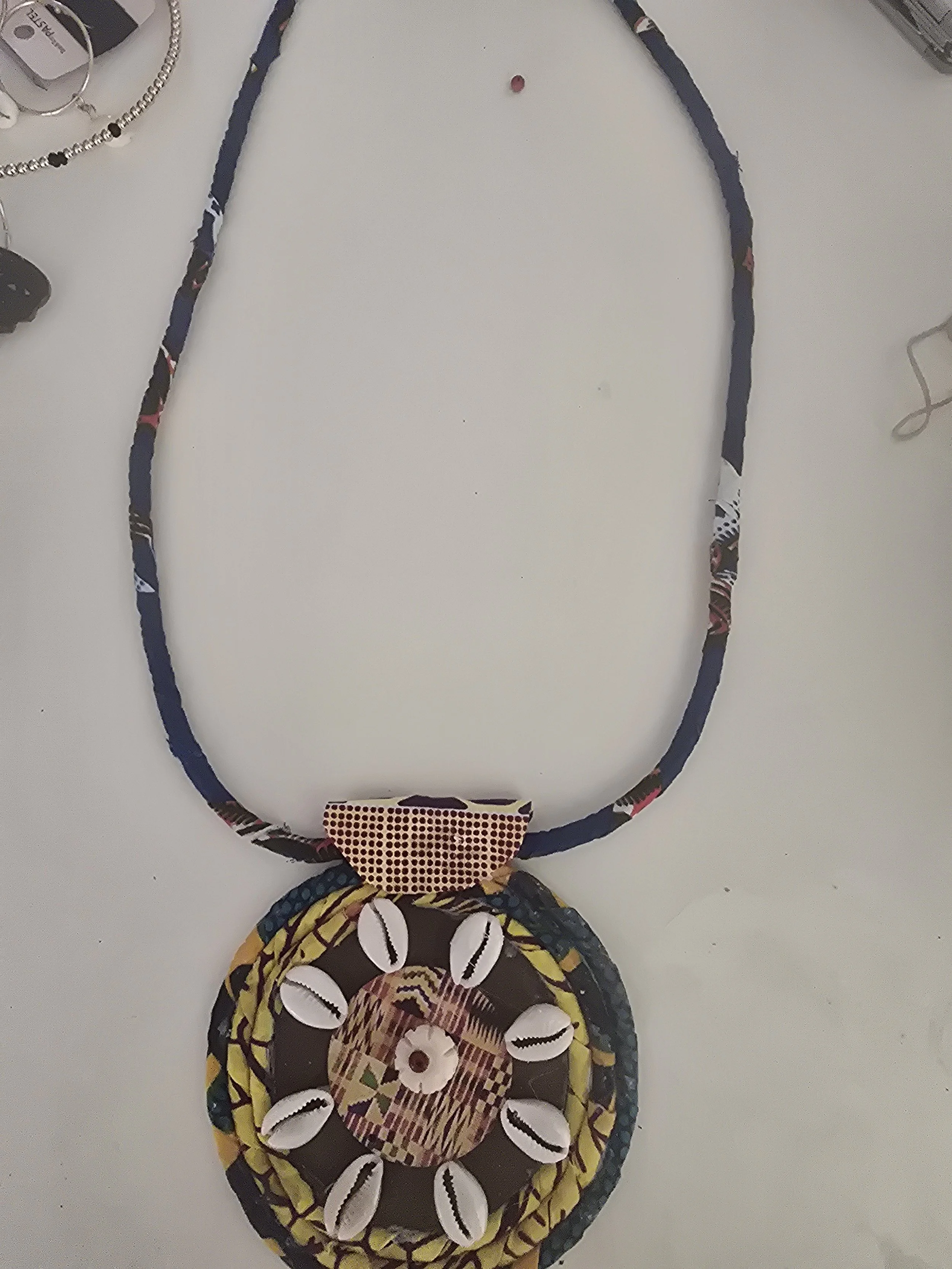Colorful handmade necklace with a large, round pendant decorated with shells and patterns, displayed on a white surface with other jewelry items nearby.