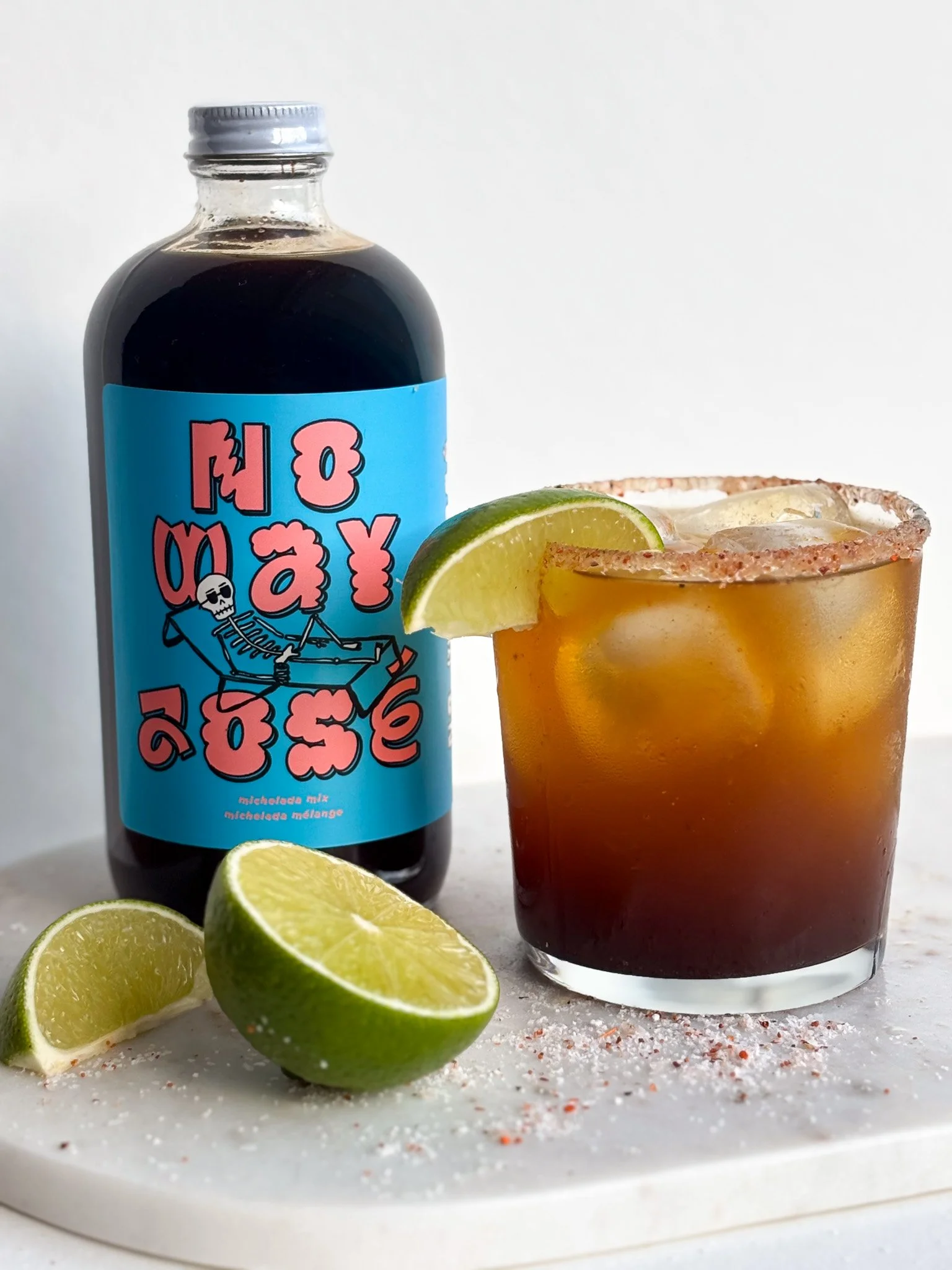 A bottle of molelada mix with a colorful label, a short glass filled with a dark cocktail, ice, a lime wedge on the rim, and lime halves with salt on the rim and on the surface, all on a white surface.