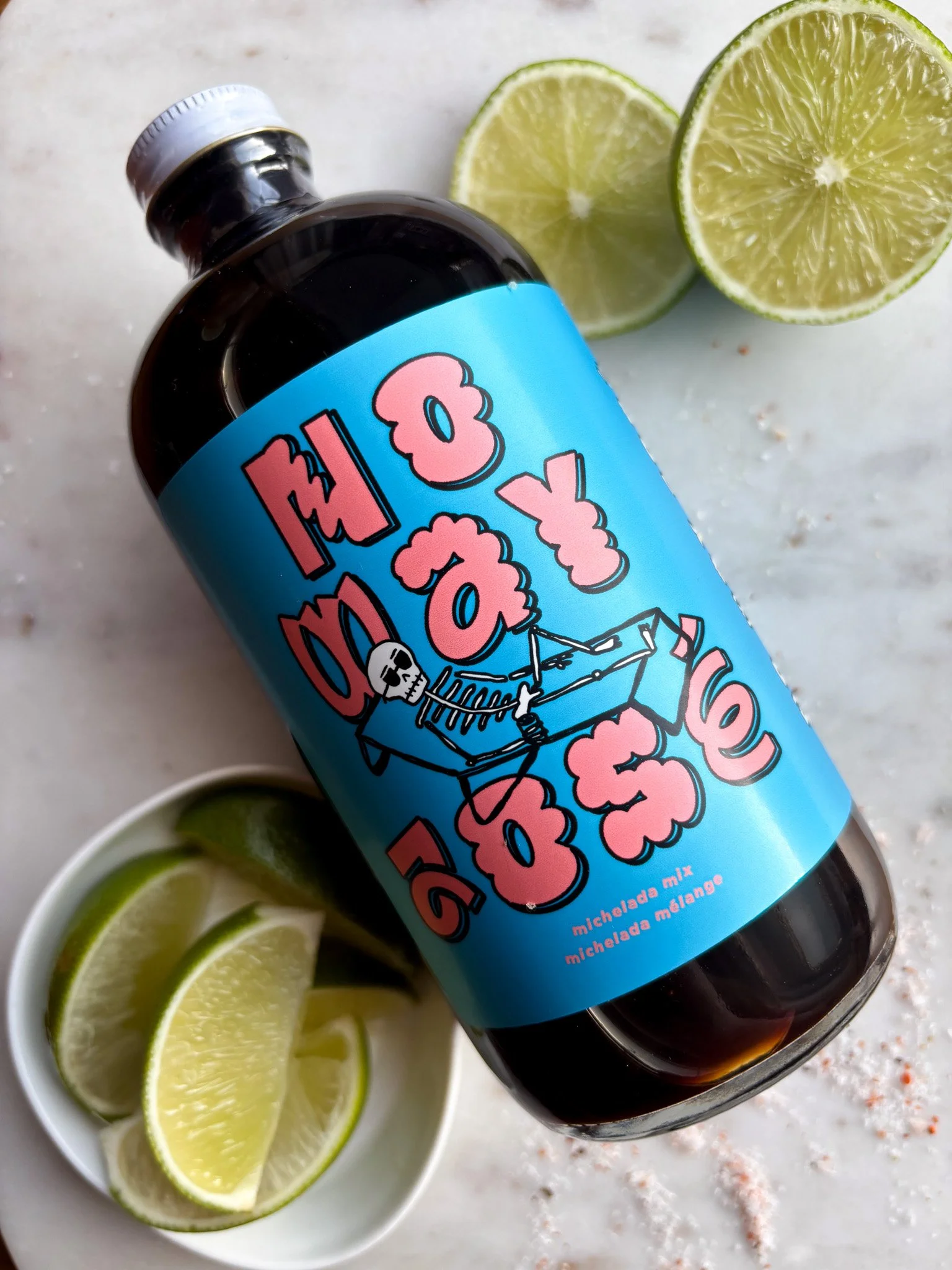 Bottle with a bright blue label reading "Hooked on Life" surrounded by lime slices and coarse salt on a white surface.