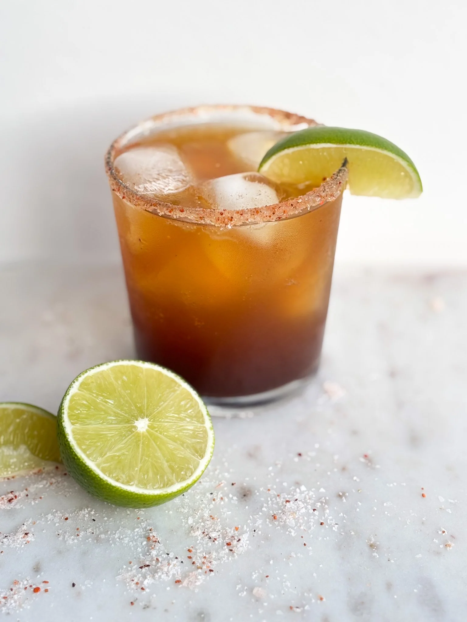 A glass of iced cocktail garnished with a lime wedge, salt rim, and lime slices on a white surface.