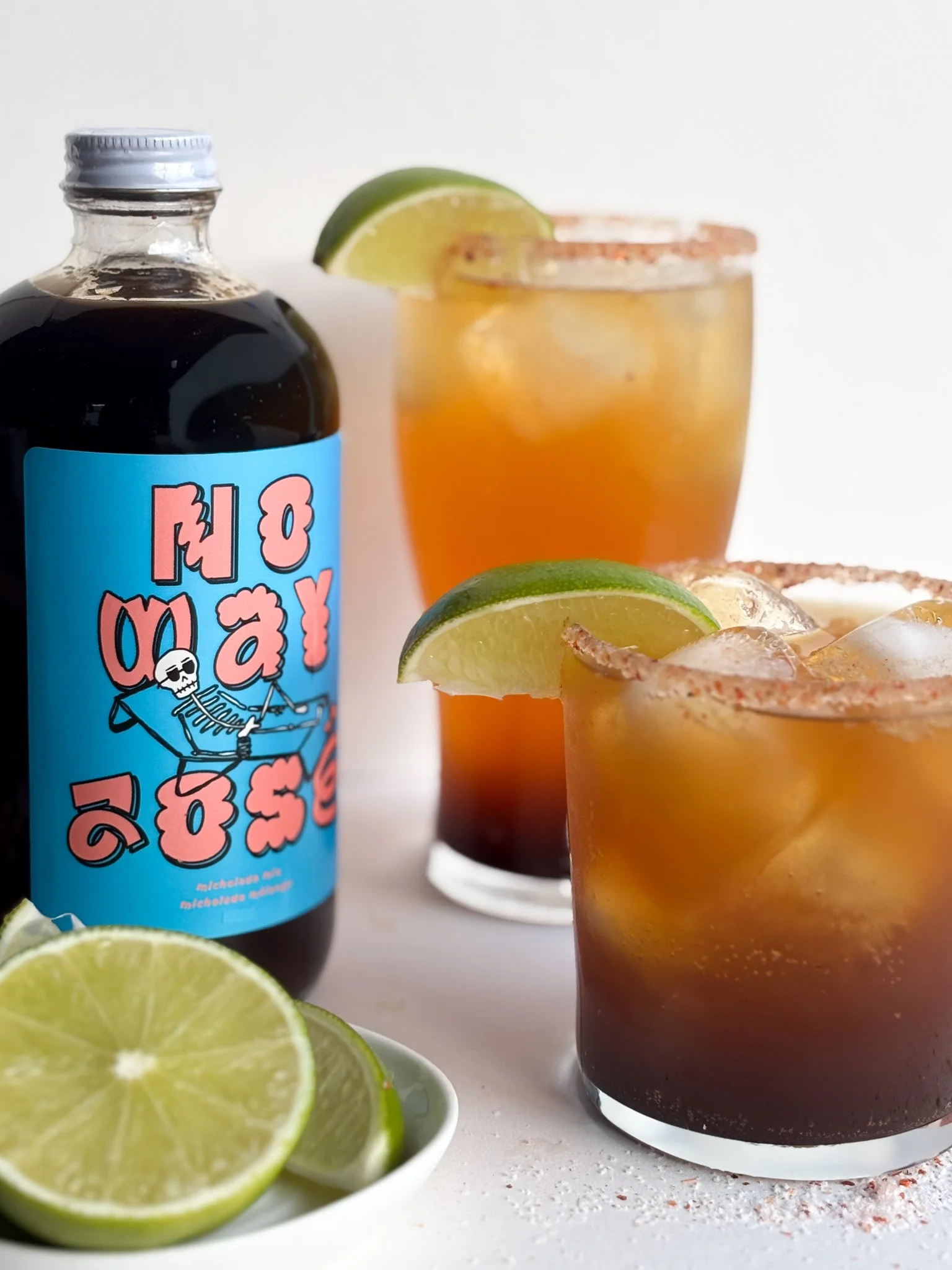 Two glasses of tequila cocktails with lime wedges, a bottle of tequila with a colorful label, and lime slices on a white surface.