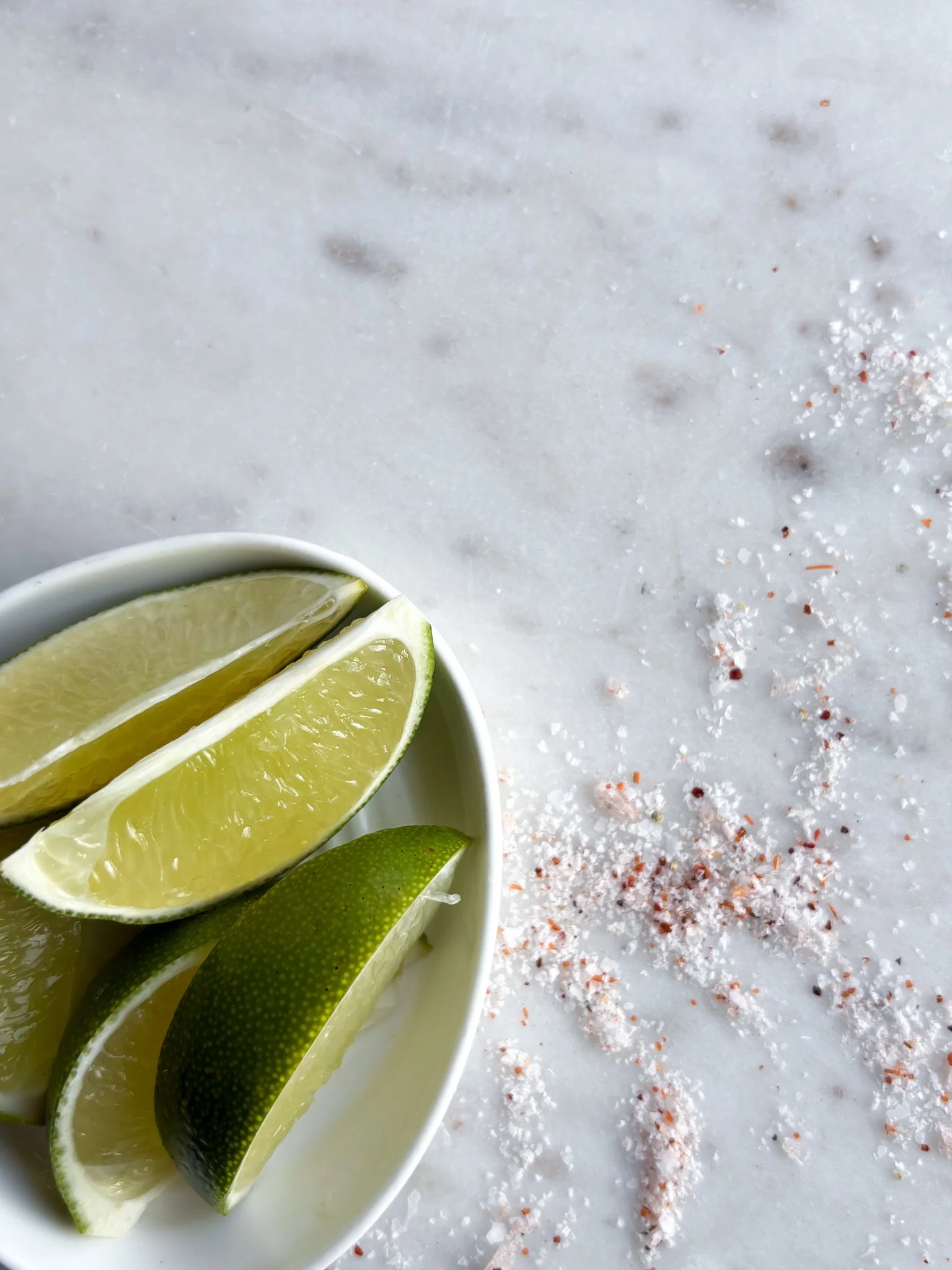 Lemon and lime wedges in a white bowl on a white marble surface, with salt and red pepper flakes scattered on the surface.