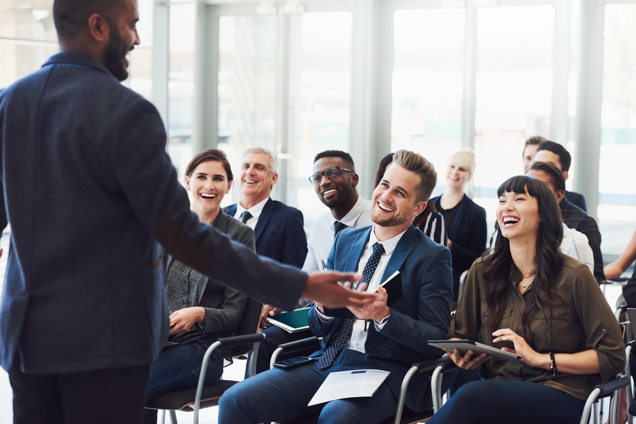A diverse group of business professionals attending a conference or seminar, smiling and laughing while listening to a speaker.
