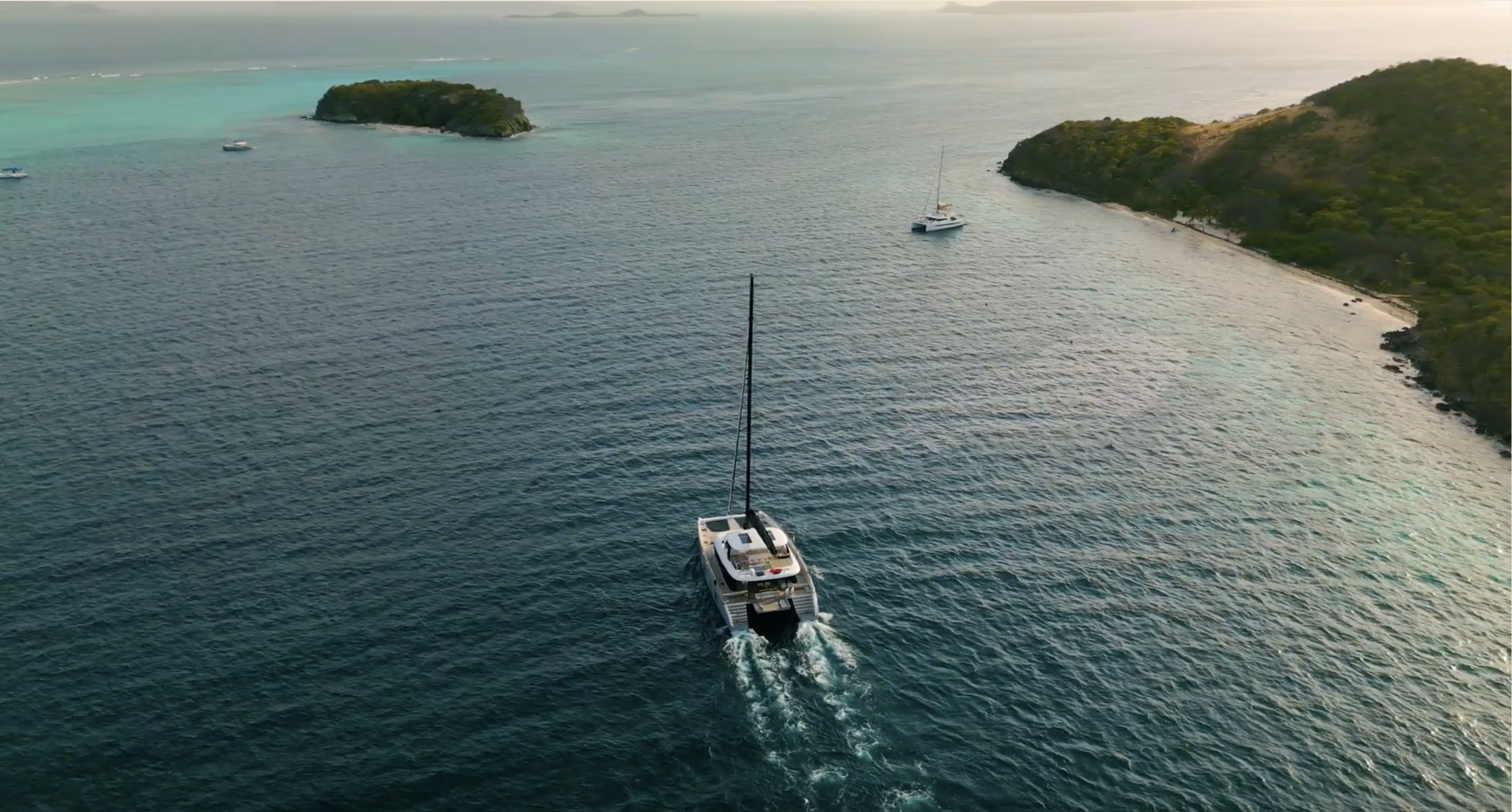 A sailboat sailing in turquoise waters near lush green islands and coastline at sunset.