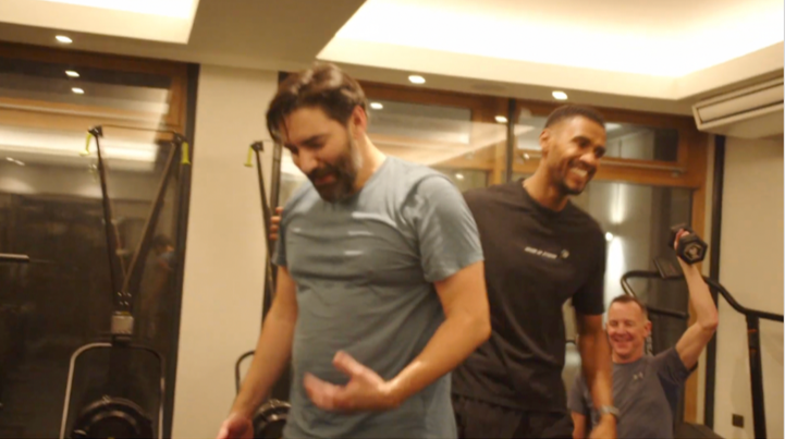 Three men smiling and sitting in a gym, one is holding a dumbbell, others are standing and talking, gym workout equipment visible in the background.
