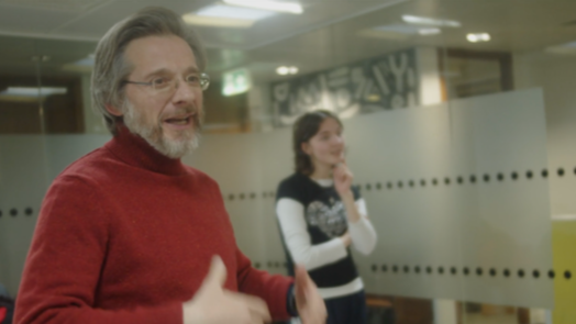 A man with gray hair and glasses, wearing a red sweater, speaking in an office setting, with a woman in the background listening.