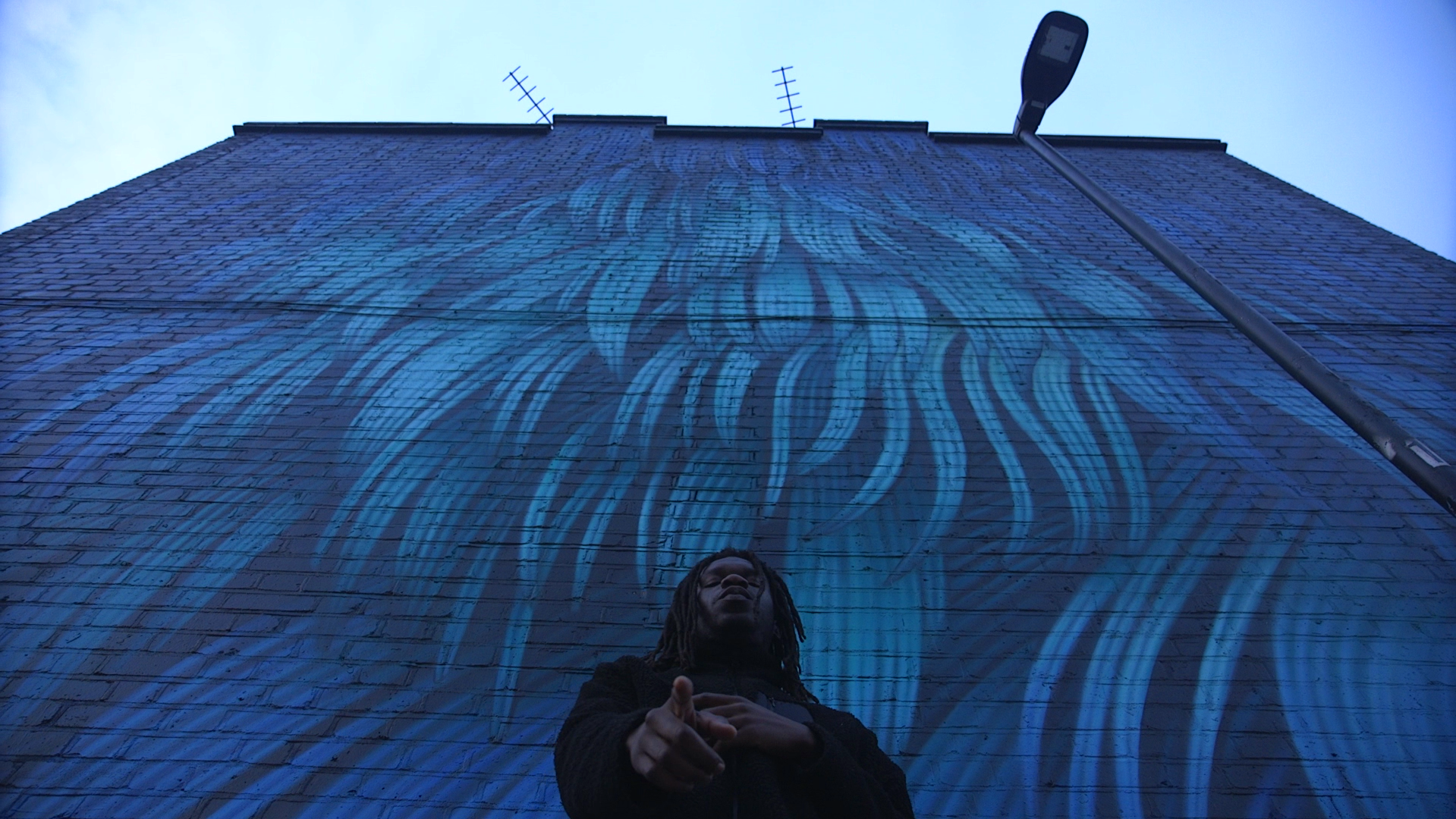 Person with dreadlocks stands in front of a tall brick building with blue flames painted on the wall. The person is looking up with their hands clasped in front of them, and a streetlamp is visible to the right.