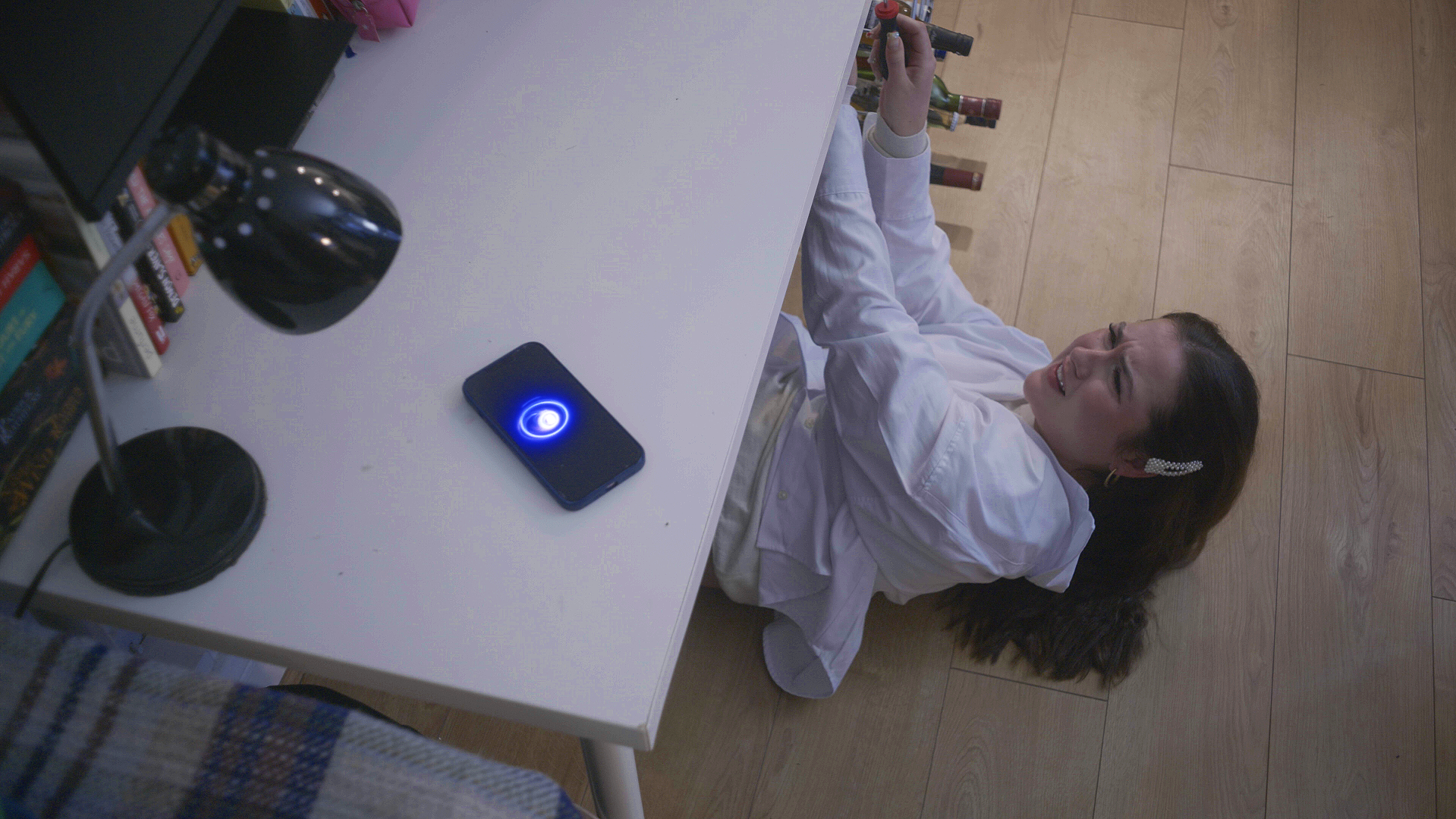 A young woman in a white shirt is lying on the wooden floor, looking up and smiling while watching a phone with an illuminated circular blue icon on the screen placed on a white desk. On the desk are various items including a black lamp, books, and a computer monitor.