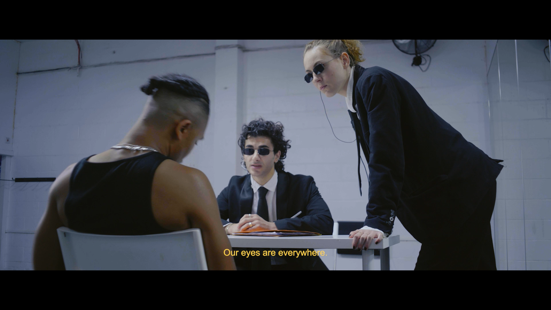 Four people in a room with white walls. One person with sunglasses and curly hair sitting at a desk, a man with sunglasses and dark suit sitting at the desk, and a woman with sunglasses and red hair leaning on the desk. The person at the desk appears to be writing or signing something. Subtitles at the bottom read, "Our eyes are everywhere."