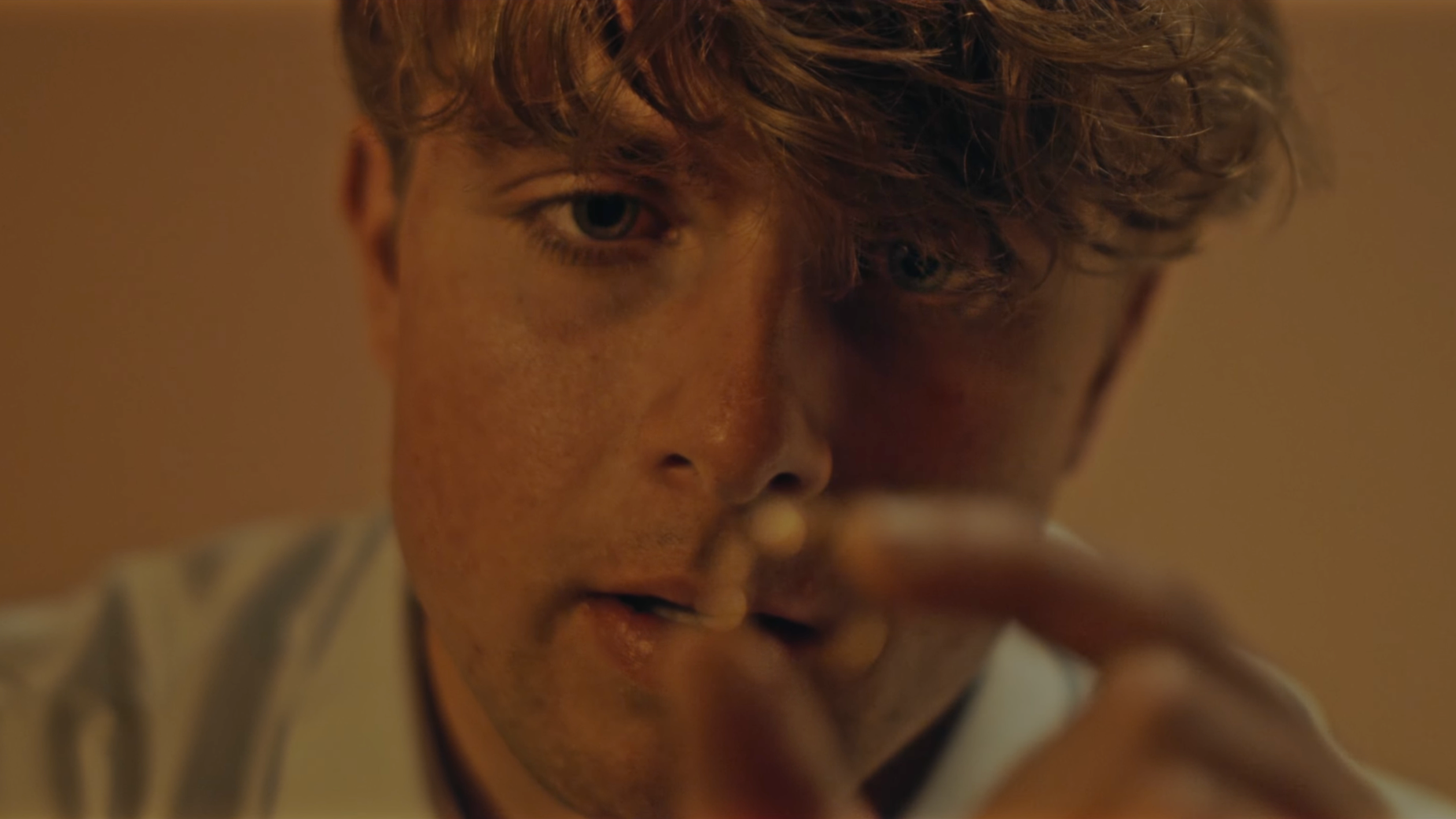 Close-up of a young man with curly brown hair, light skin, and freckles, looking intensely at a camera, with part of his face and hand visible.