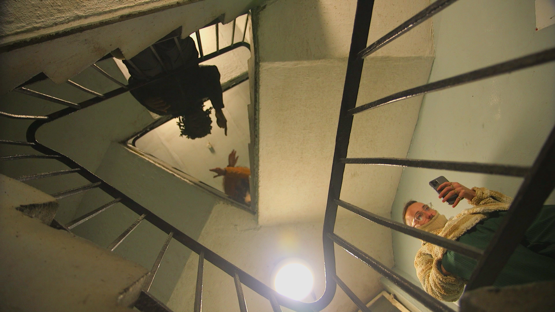People looking down from the top of a staircase with a square mirror reflecting the scene below. One person is taking a photo with a smartphone, and another is reaching out their hand.